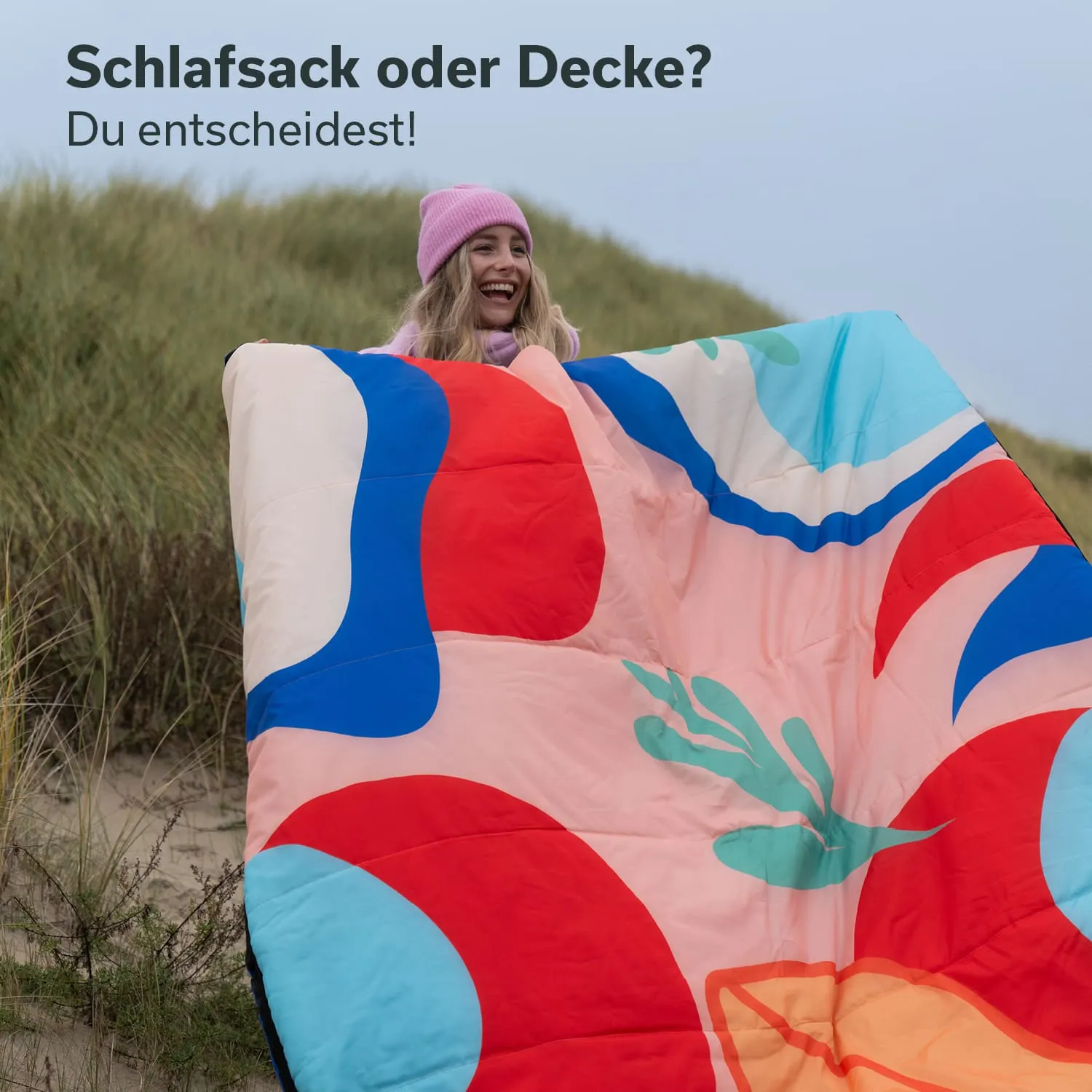 Frau mit blonden Haaren hält einen bunten Schlafsack oder eine Decke vor sich, steht in einer grasbewachsenen Umgebung.