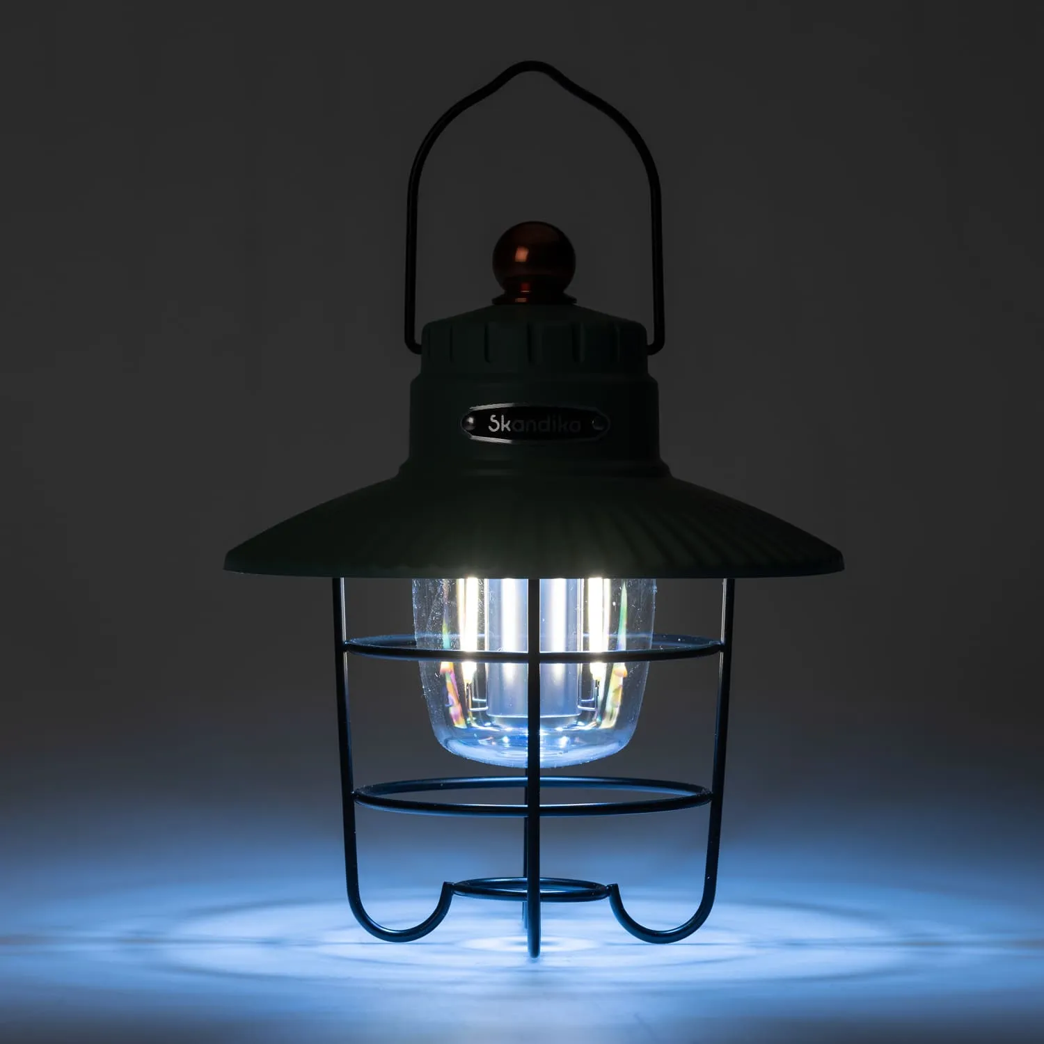 Öllampe mit grünem Gehäuse und Glaskörper, umgeben von einem schwarzen Metallgitter, leuchtet in schwachem blauem Licht.