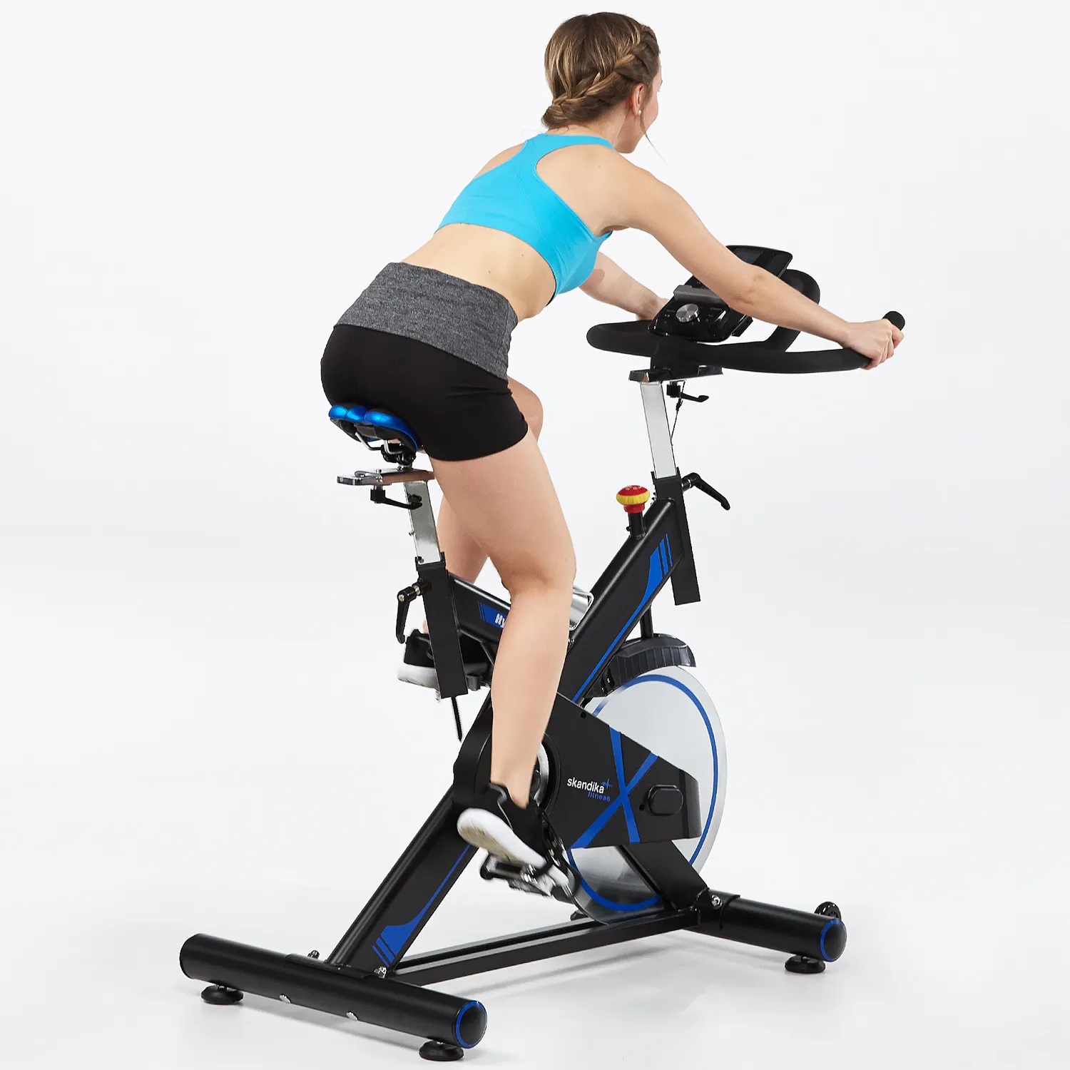 Person auf einem Fitness-Bike in sportlicher Kleidung, von hinten sichtbar, in neutralem Raum mit hellen Wänden.