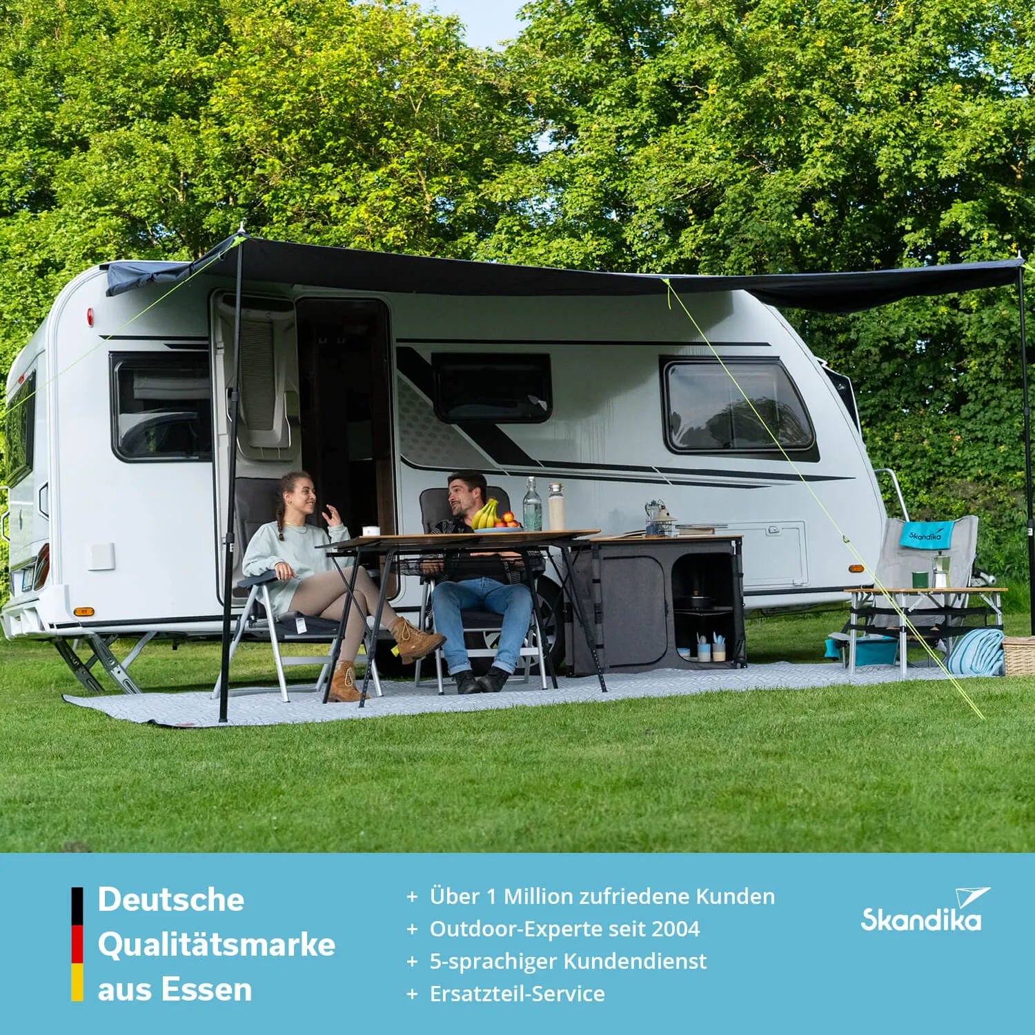 Wohnmobil auf Rasen, zwei Personen an einem Tisch mit Obst und Getränken, Sonnenschutzdach, Bäume im Hintergrund.