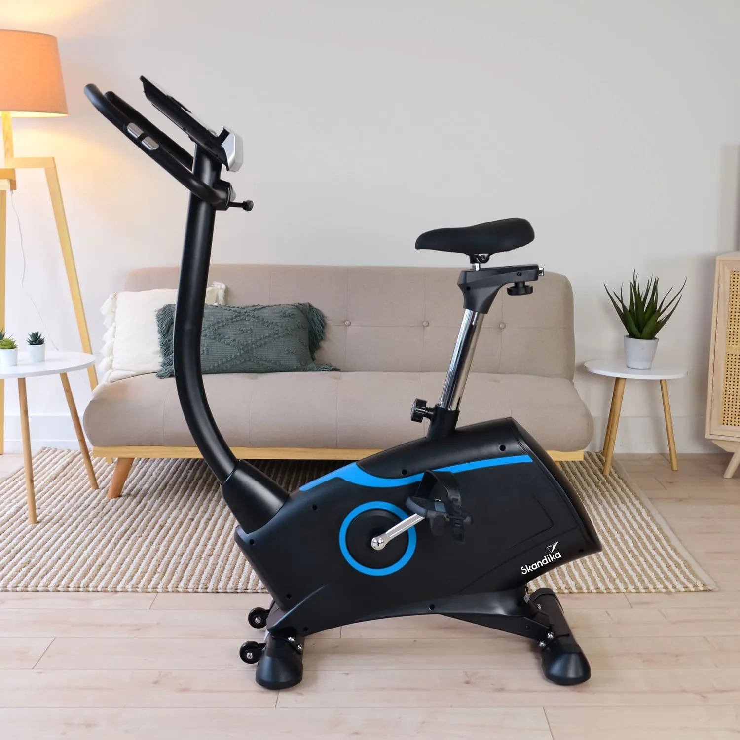 Ein Fahrradergometer mit schwarzem Gehäuse und blauen Akzenten steht in einem hellen Raum. Im Hintergrund befindet sich eine Couch und eine Pflanze.
