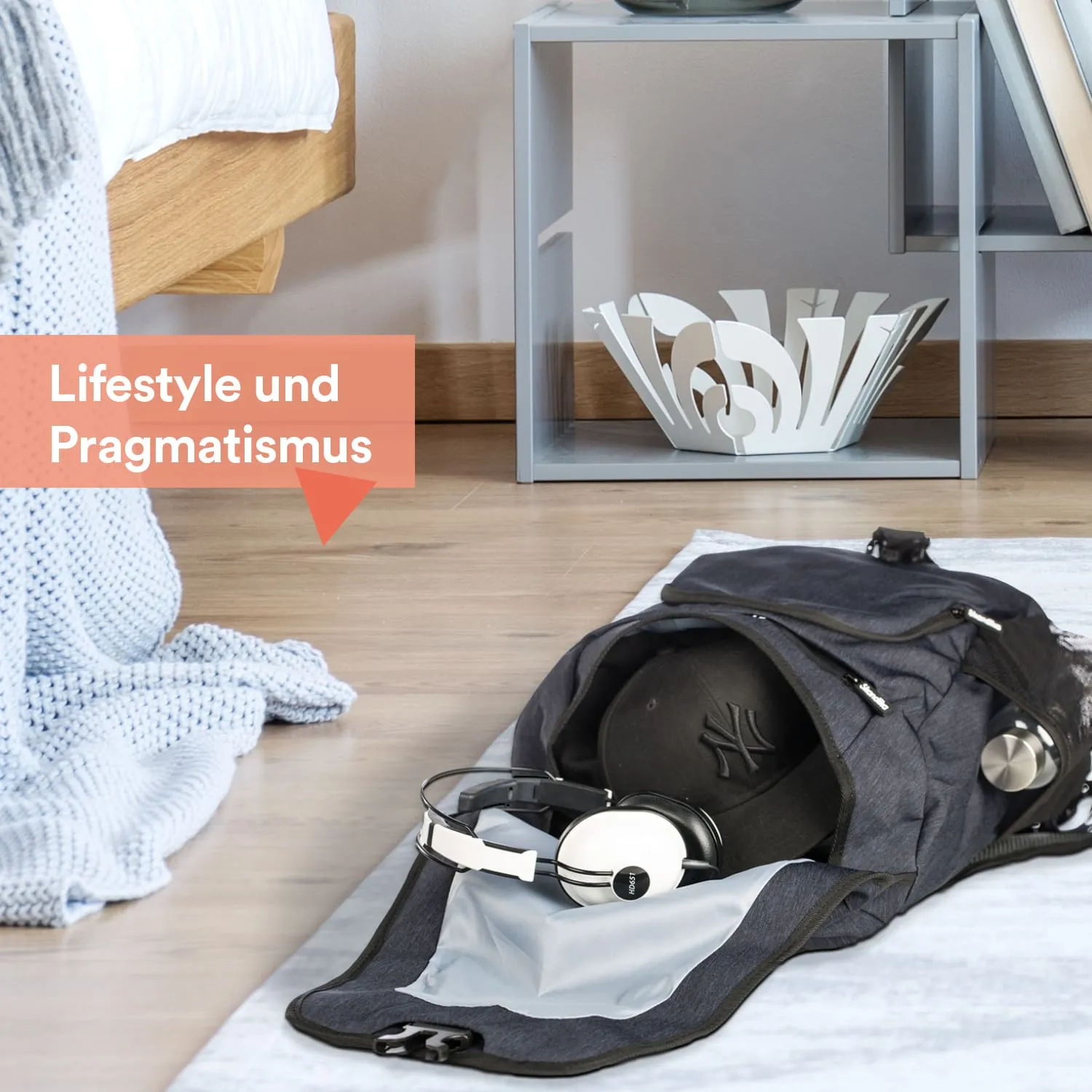 Schwarzer Rucksack mit Kopfhörern und einer Kappe auf einem Holzboden, daneben eine graue Ablage und eine Decke.