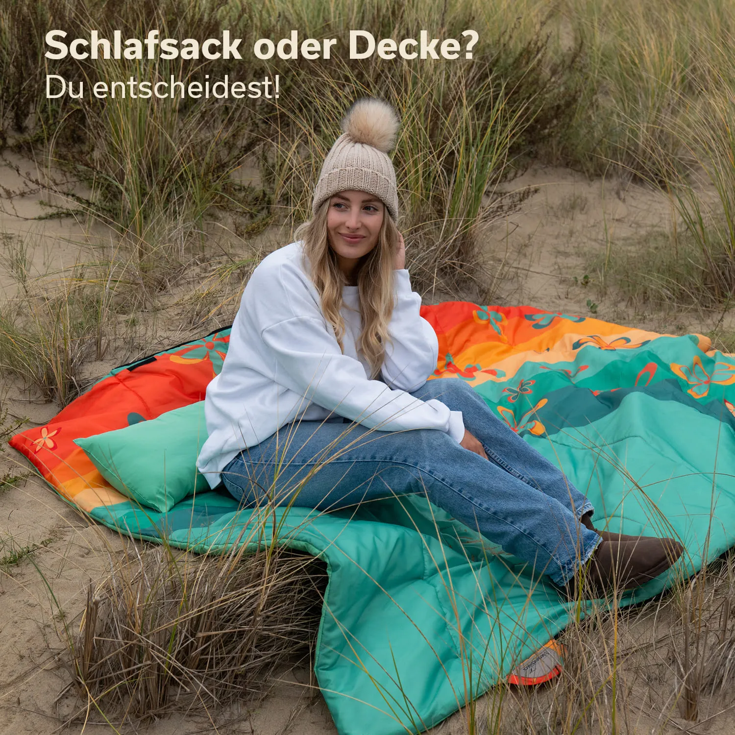 Eine Person sitzt auf einer bunten Decke im Freien, umgeben von hohem Gras. Die Person trägt einen Pullover und eine Mütze mit Pompons. Es sind weitere bunte Kissen sichtbar.