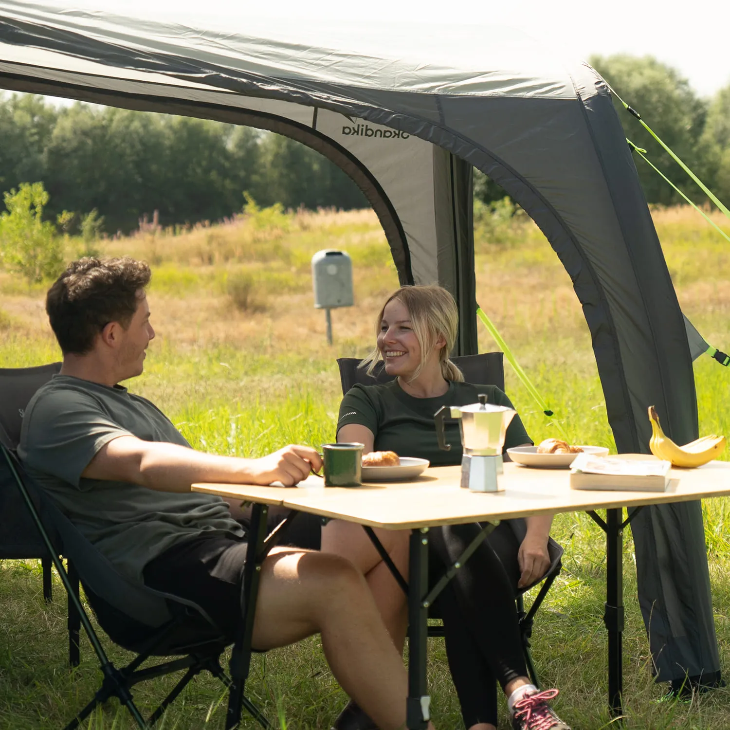 Zwei Personen sitzen unter einem Zelt auf Campingstühlen an einem Tisch. Auf dem Tisch befinden sich Geschirr mit Gebäck und eine Kaffeekanne. Im Hintergrund Wiese und Bäume.