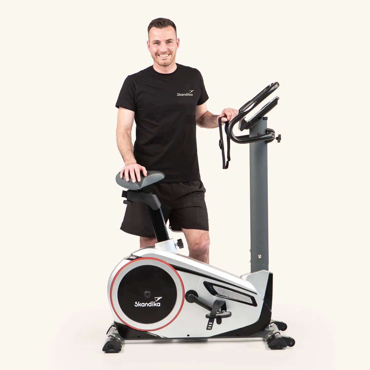 Ein Indoor-Fahrrad steht vor einem hellen Hintergrund. Eine Person lächelt, steht neben dem Fahrrad und hält einen Griff in der Hand. Die Person trägt ein schwarzes T-Shirt und kurze Hosen. Auf dem Fahrrad ist das Logo „Skandika“ sichtbar.