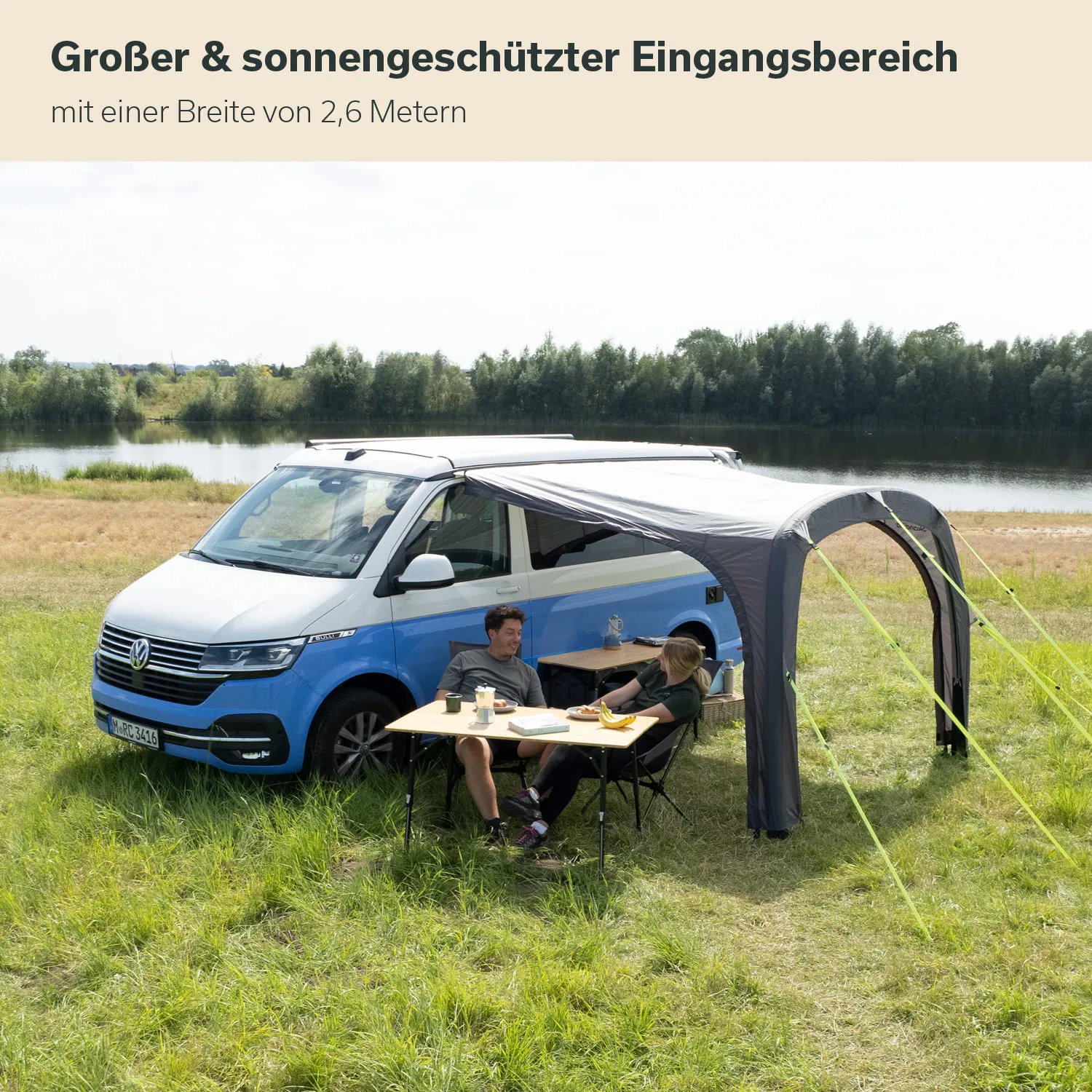 Ein blauer Campingbus steht auf einer Wiese am Wasser. Unter einem Sonnenschutz sitzen zwei Personen an einem Tisch.