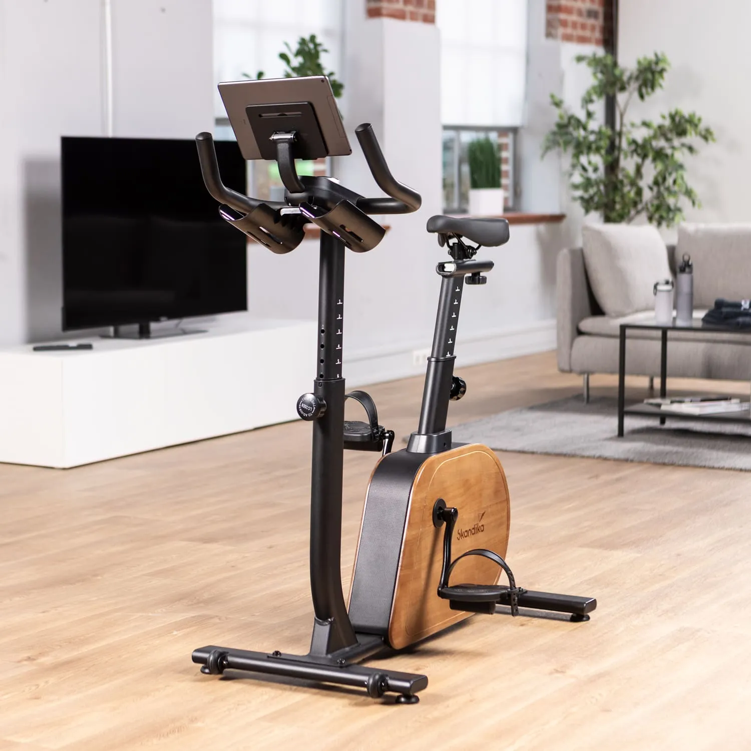 Ein stationäres Fahrrad in einem modernen Raum. Das Rad hat einen schwarzen Rahmen und eine Holzunterlage, mit einem Bildschirm an der Vorderseite. Im Hintergrund steht ein Fernseher und ein Sofa.