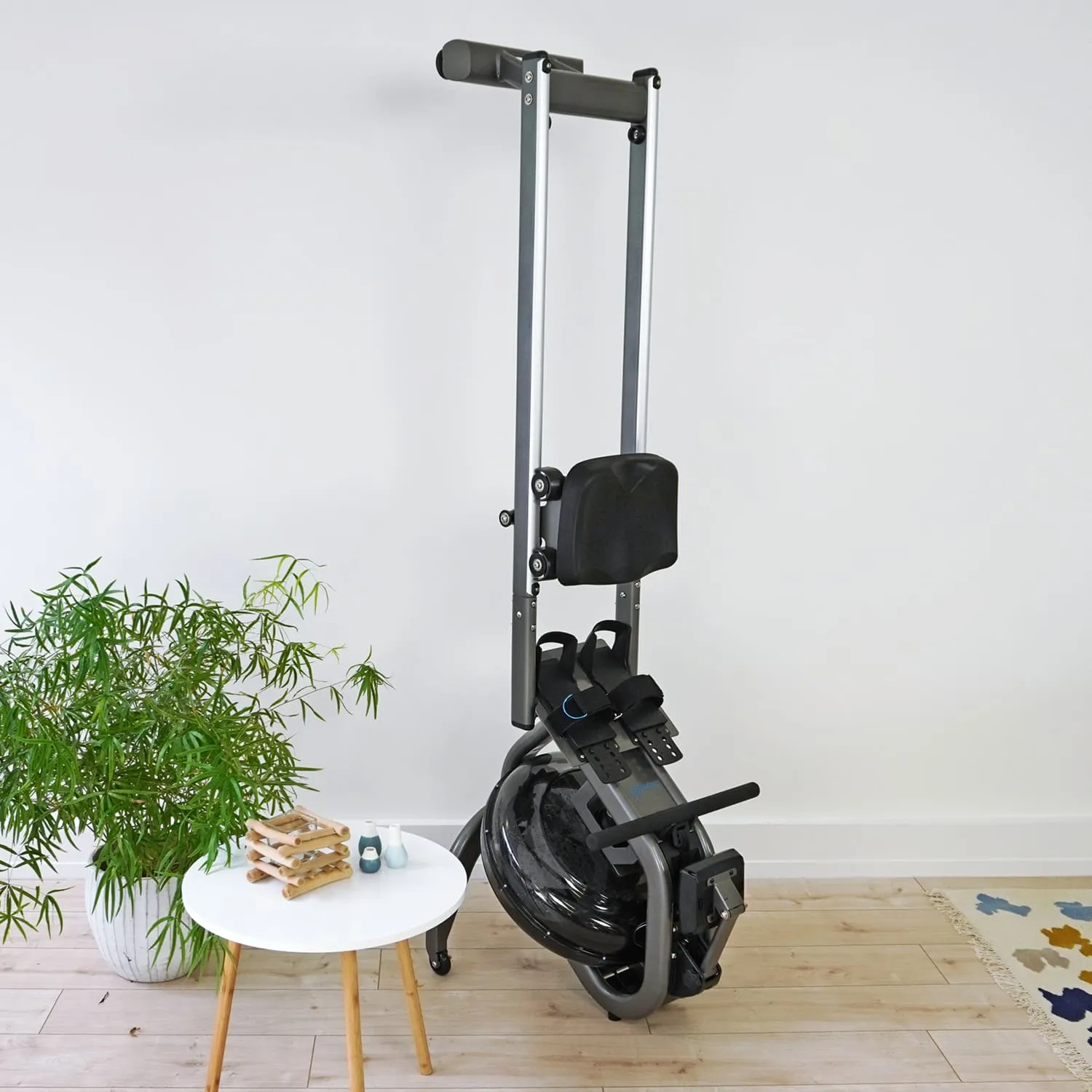 Rudergerät mit schwarzem Sitz und Pedalen, neben einem kleinen runden Tisch und einer Pflanzen in einem hellen Raum.
