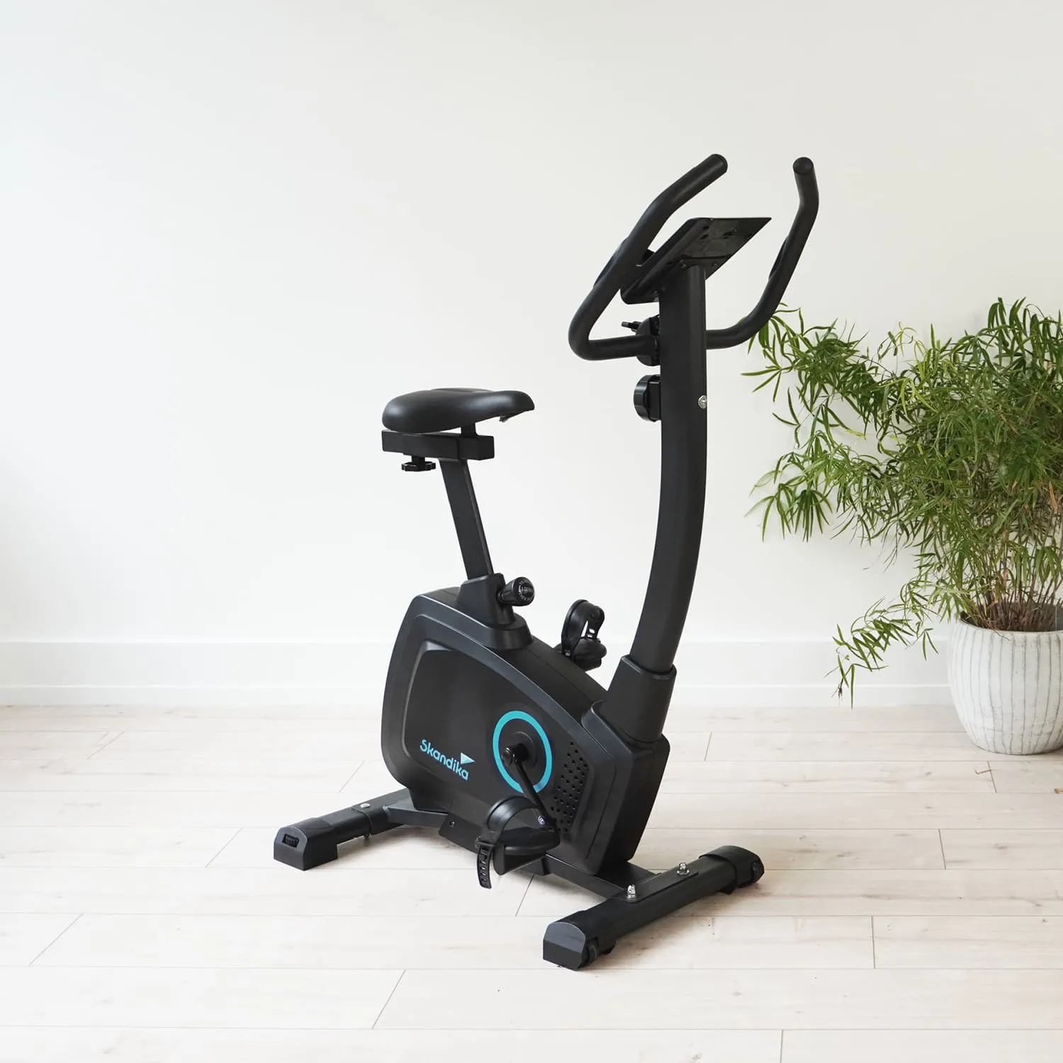 Ergometer in Schwarz mit einem blauen Detail, stehend auf einem hellen Holzboden neben einer Pflanze in einem Topf.