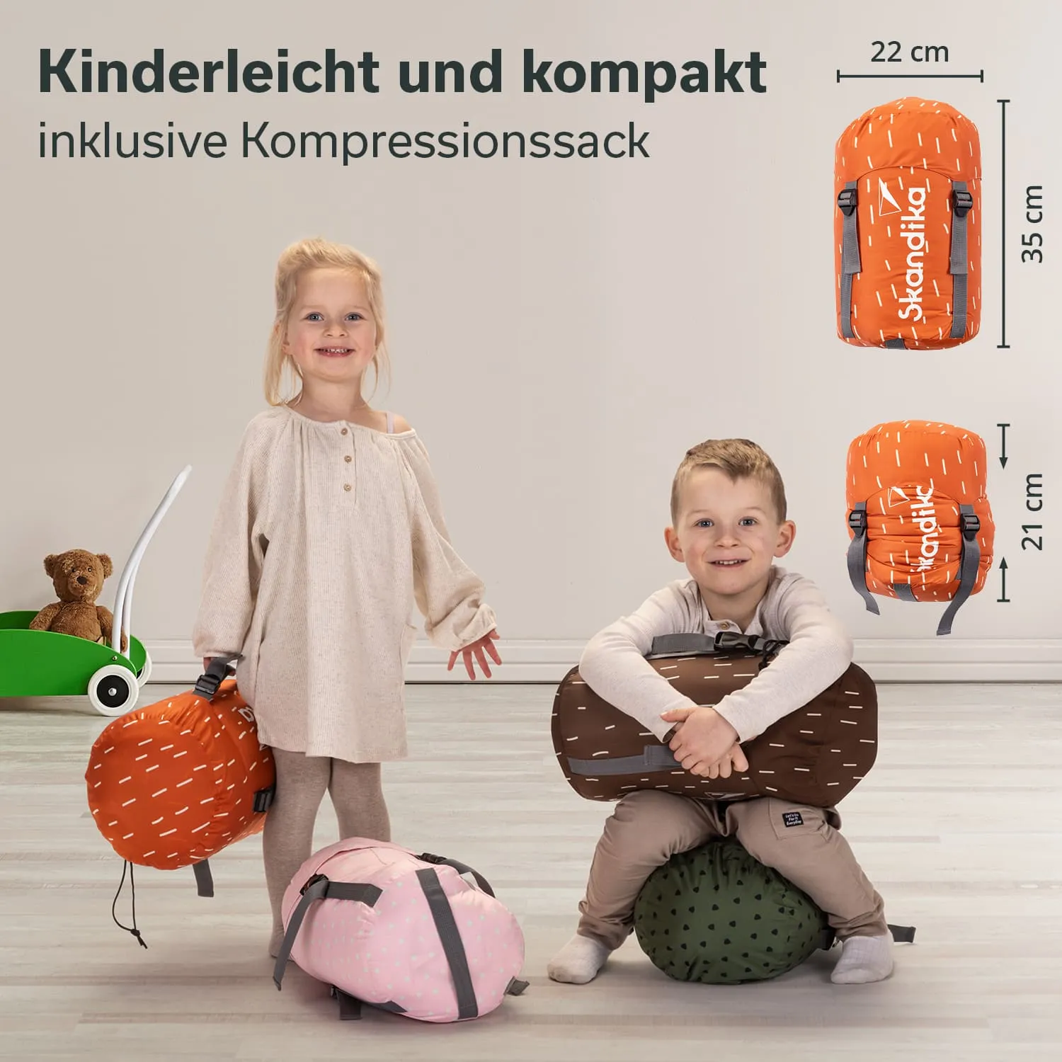 Zwei Kinder stehen in einem hellen Raum. Das Mädchen trägt ein beiges Oberteil und hält einen orangefarbenen Kompressionssack, während der Junge auf einem grünen Kissen sitzt und einen braunen Sack umarmt. Ein rosa Sack liegt auf dem Boden.