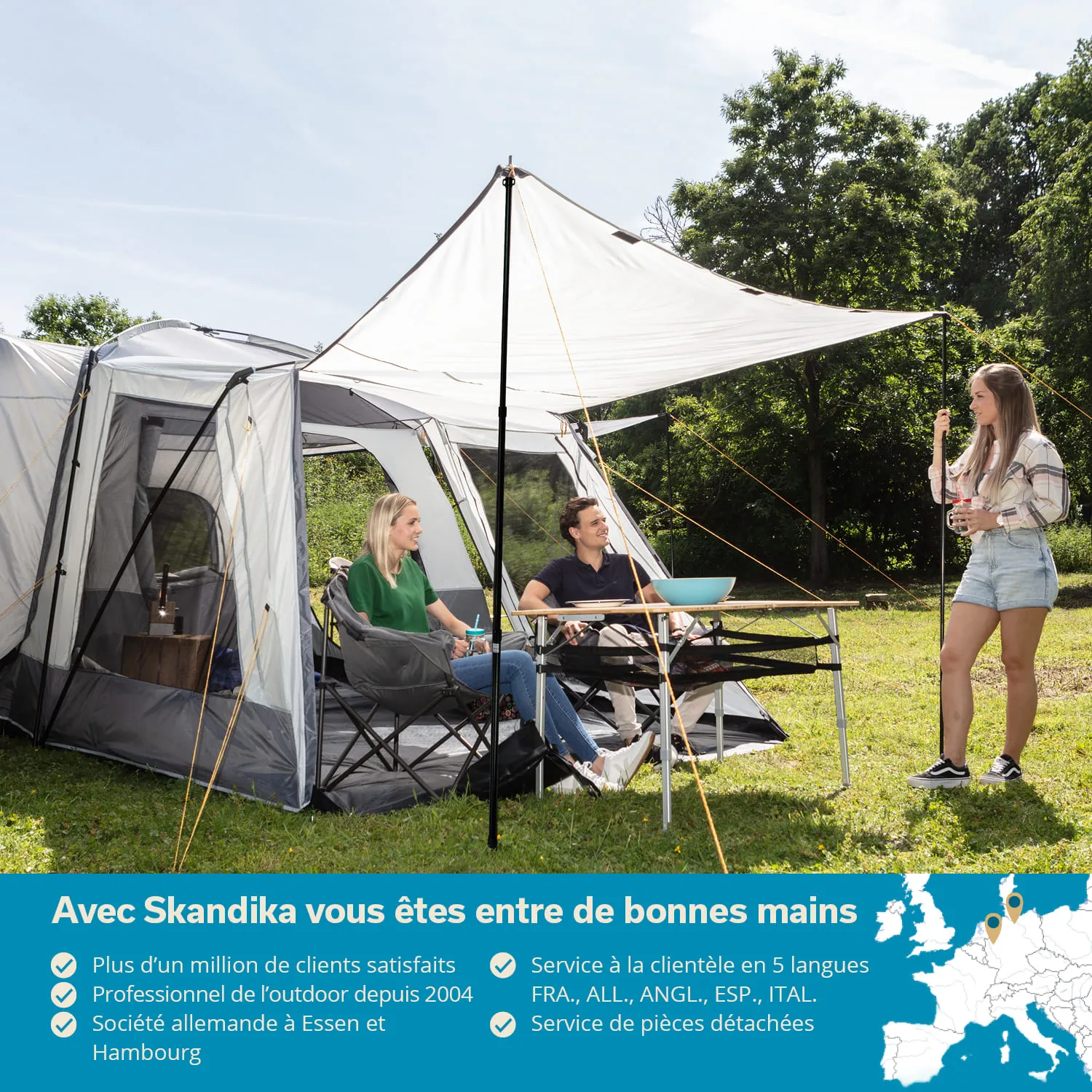 Drei Personen sitzen in Campingstühlen unter einem Zelt mit weißem Sonnenschutz. Eine Person steht und spricht. Hintergrund mit Bäumen.