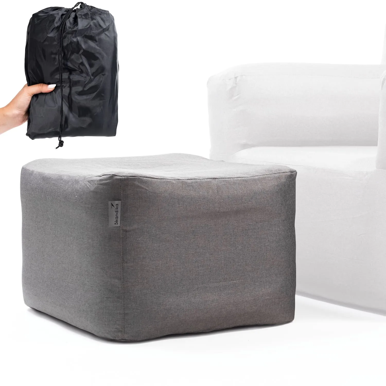 Grauer Pouf neben einem weißen Sessel. Eine Person hält eine schwarze, gefaltete Tasche.