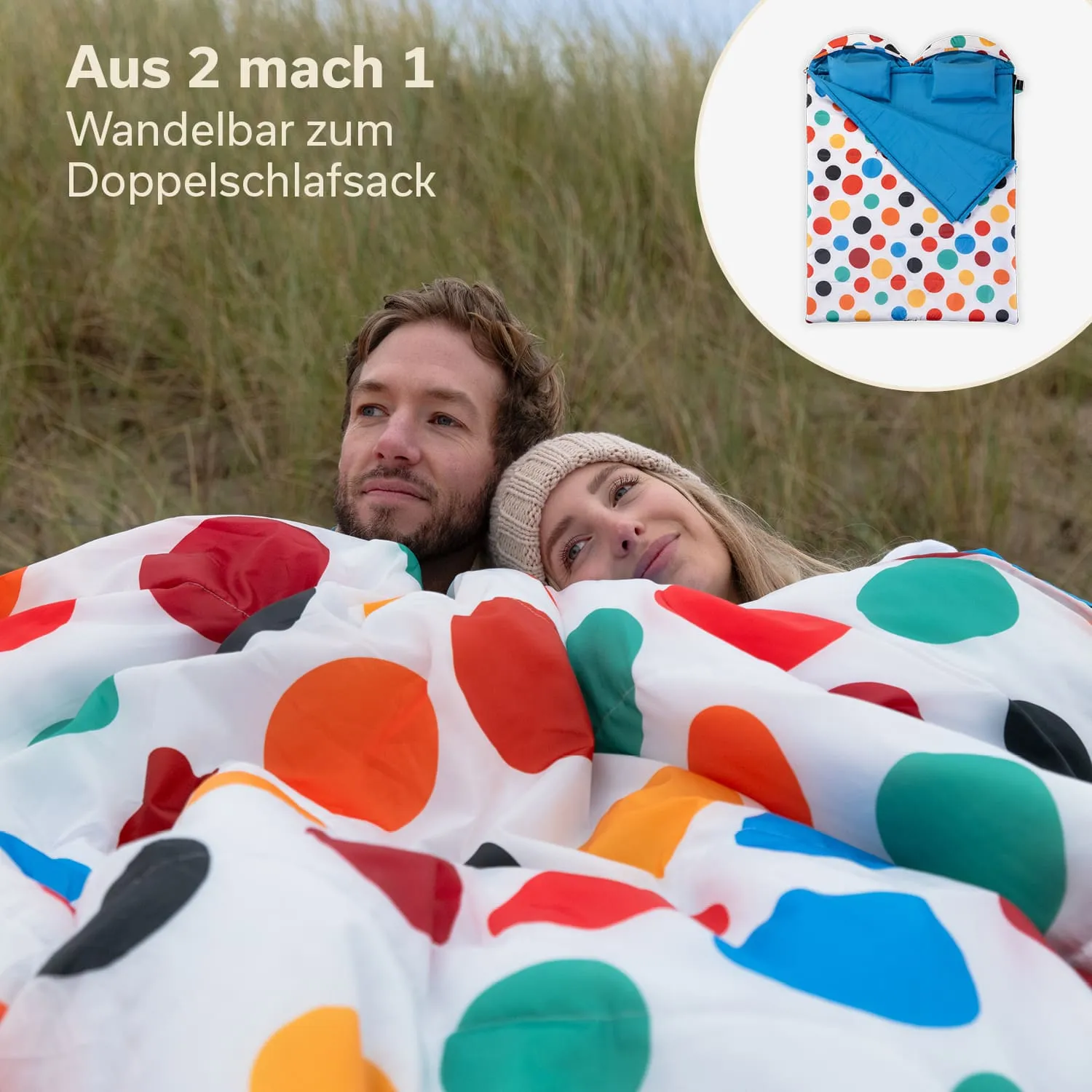 Zwei Personen liegen unter einem farbigen Schlafsack mit großen Punkten. Im Hintergrund sind Dünen zu sehen.