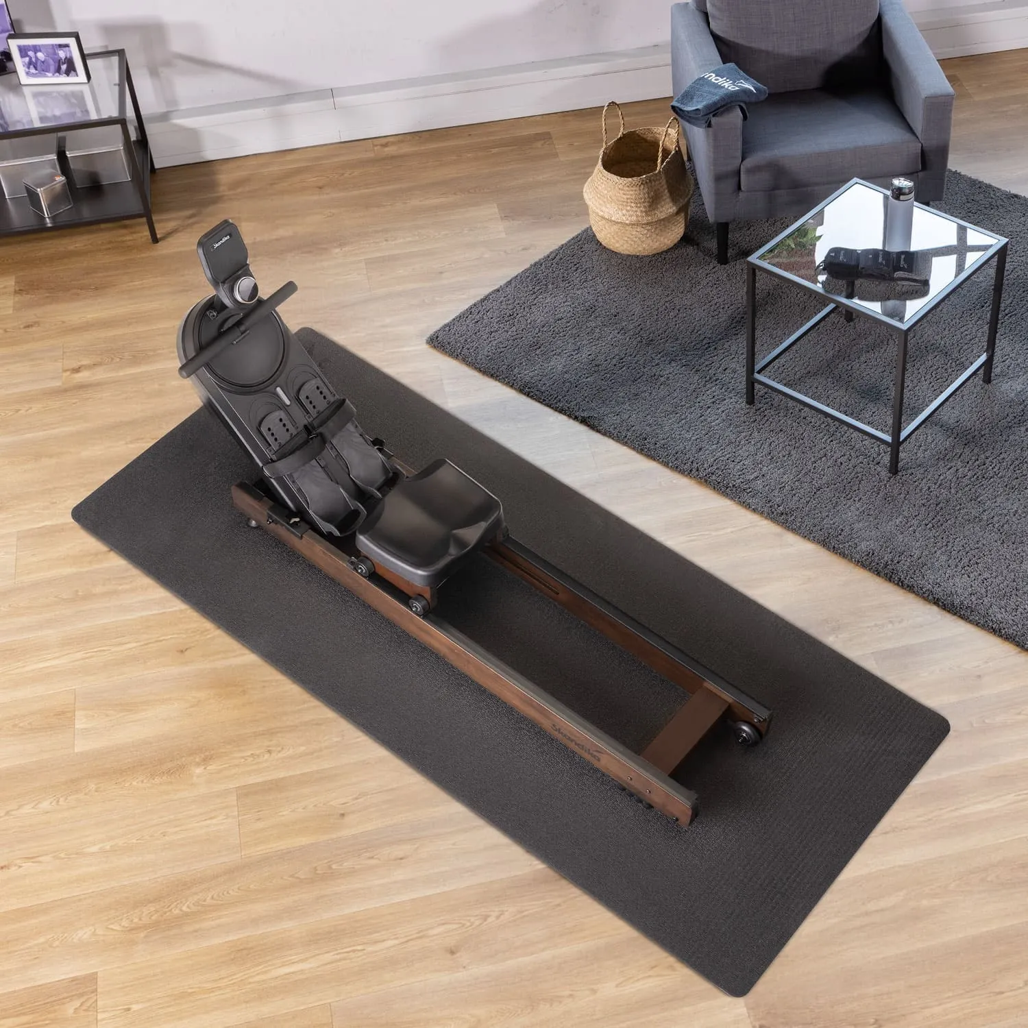 Rudergerät auf einer dunklen Matte, Holzrahmen, schwarzer Sitz, daneben ein grauer Sessel und Couchtisch mit Accessoires.