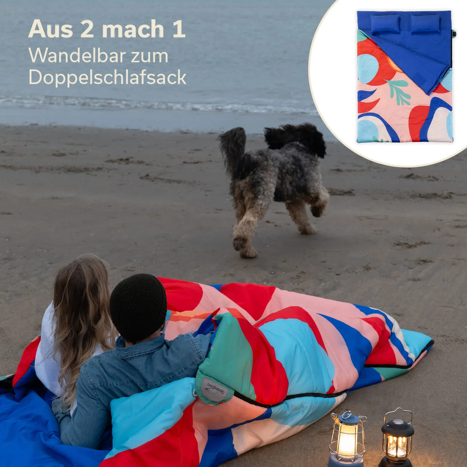 Paar sitzt auf einem Strand mit buntem Schlafsack. Ein Hund läuft im Hintergrund. Nachtbeleuchtung durch Laternen sichtbar.