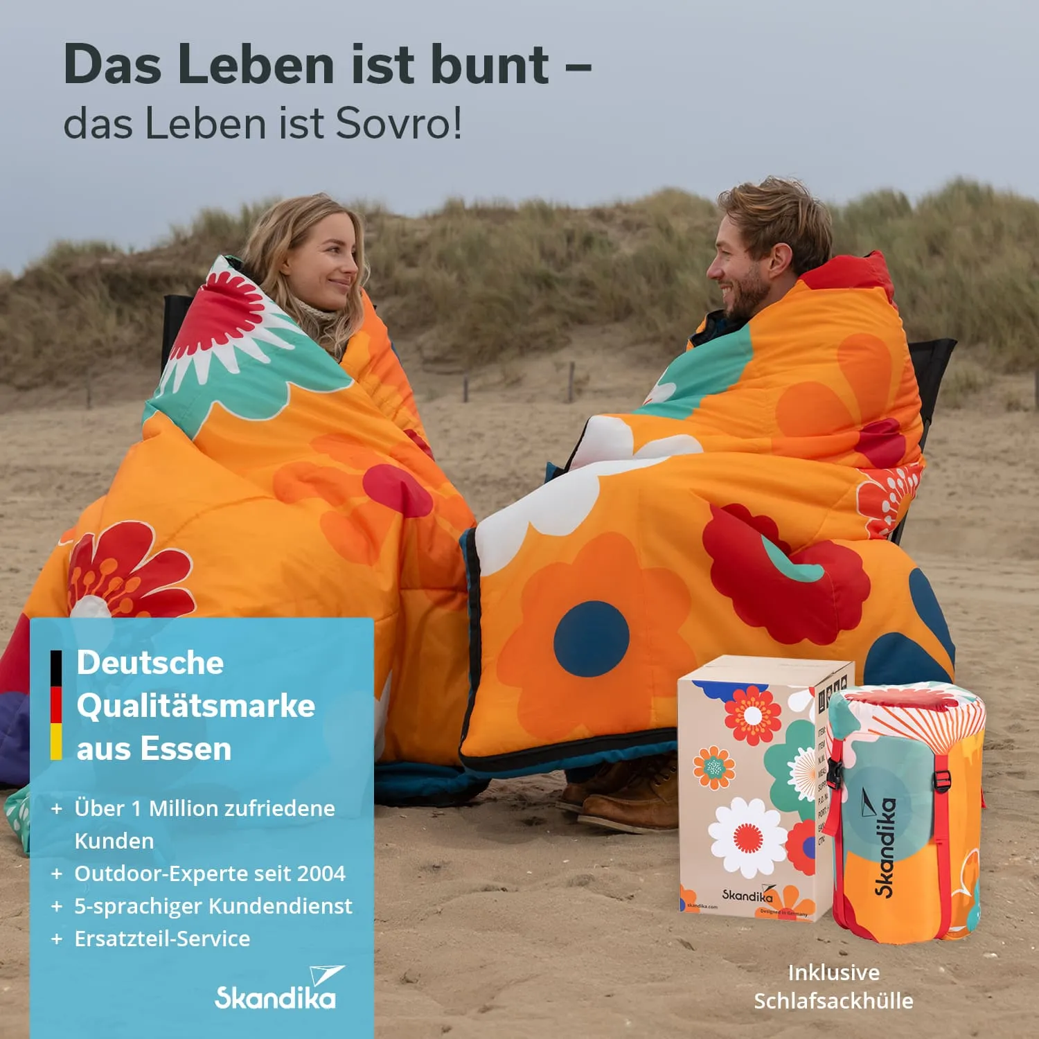 Zwei Personen sitzen im Freien am Strand, eingehüllt in bunte Schlafsäcke mit Blumenmotiven. Hinter ihnen sind Sanddünen sichtbar. Rechts steht eine Verpackung mit einem ähnlichen Design.