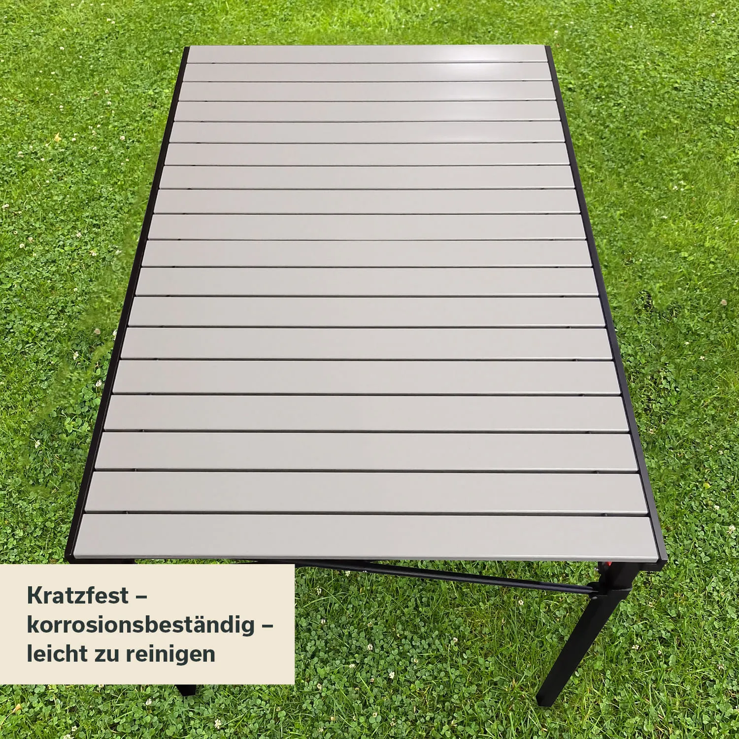 Ein grauer Tisch mit horizontalen Laminatleisten auf grünem Gras. Links unten steht ein Schild mit Produktinformationen.