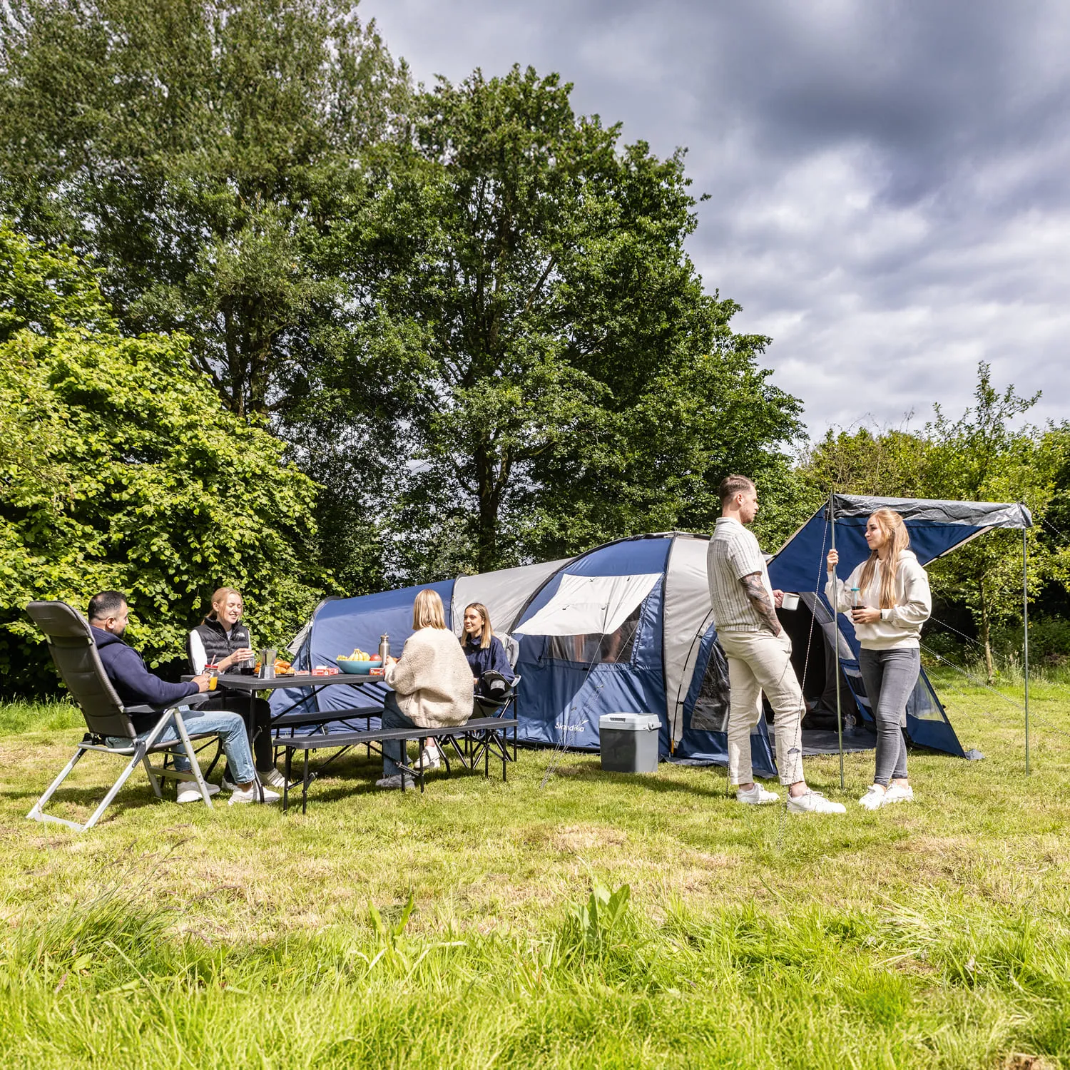 Gruppe von fünf Personen sitzt und steht um ein Zelt auf einer Wiese, umgeben von Bäumen. Ein Tisch ist gedeckt.