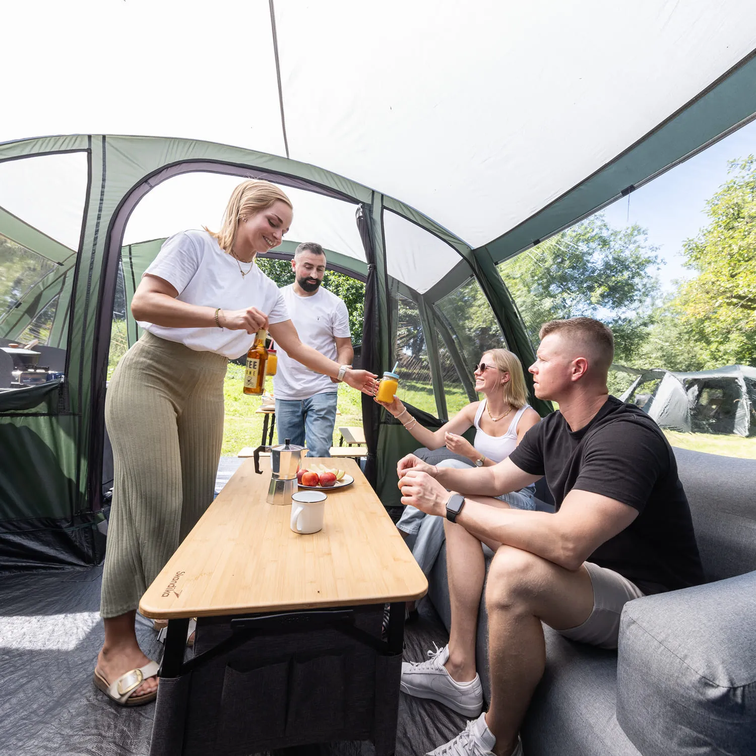 Personen sitzen innerhalb eines Zeltes an einem Tisch. Eine Person reicht eine Flasche einer anderen Person, während Obst auf dem Tisch steht.