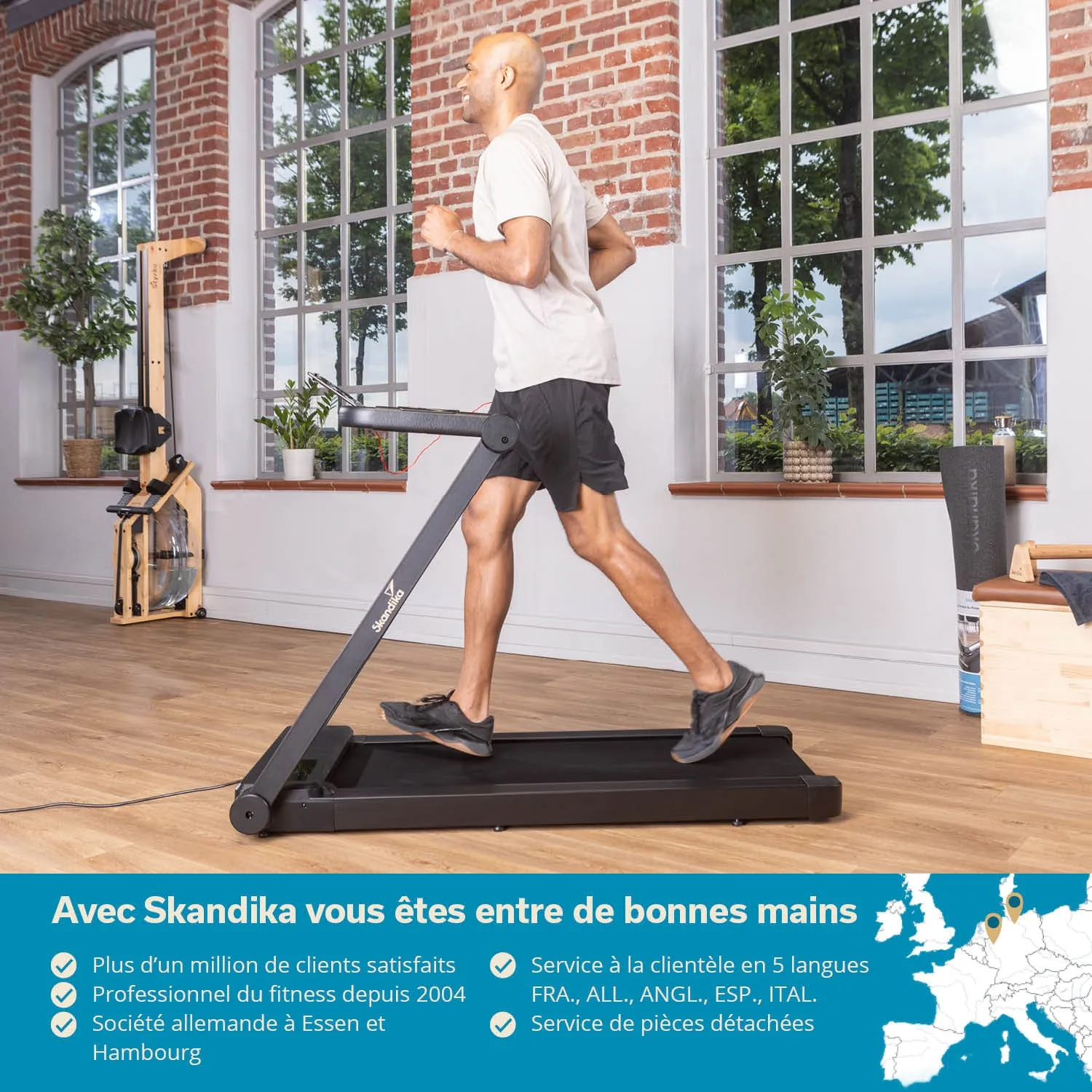 Mann joggt auf einem Laufband in einem hellen Raum mit großen Fenstern und Holzfußboden. Pflanzen sind sichtbar.