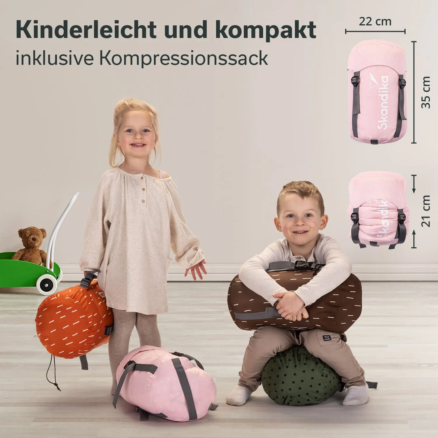 Zwei Kinder stehen auf einem hellen Boden neben bunten Taschen in verschiedenen Größen. Im Hintergrund steht ein Spielzeugwagen mit einem Stofftier.