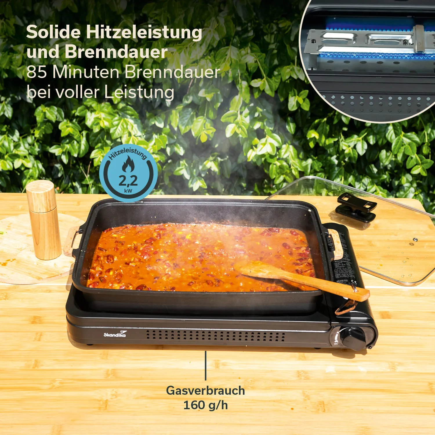 Gusseisenpfanne mit dampfendem Eintopf auf einem Tisch aus Holz. Im Hintergrund grüne Pflanzen. Es stehen Angaben zu Hitzeleistung und Gasverbrauch.