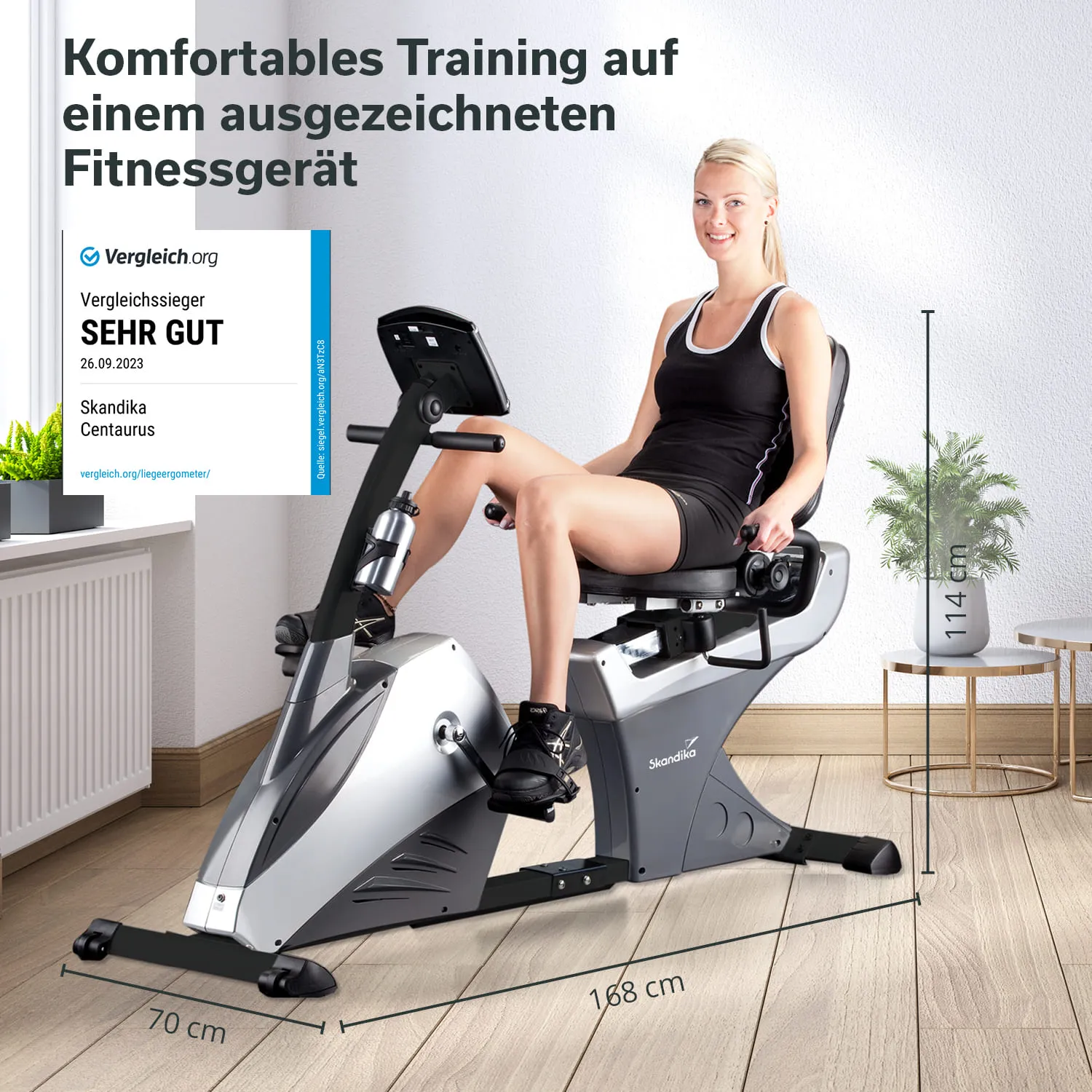 Fitnessgerät in einem Raum, eine Person sitzt darauf. An der Wand steht eine Pflanze, Holzfußboden ist sichtbar.