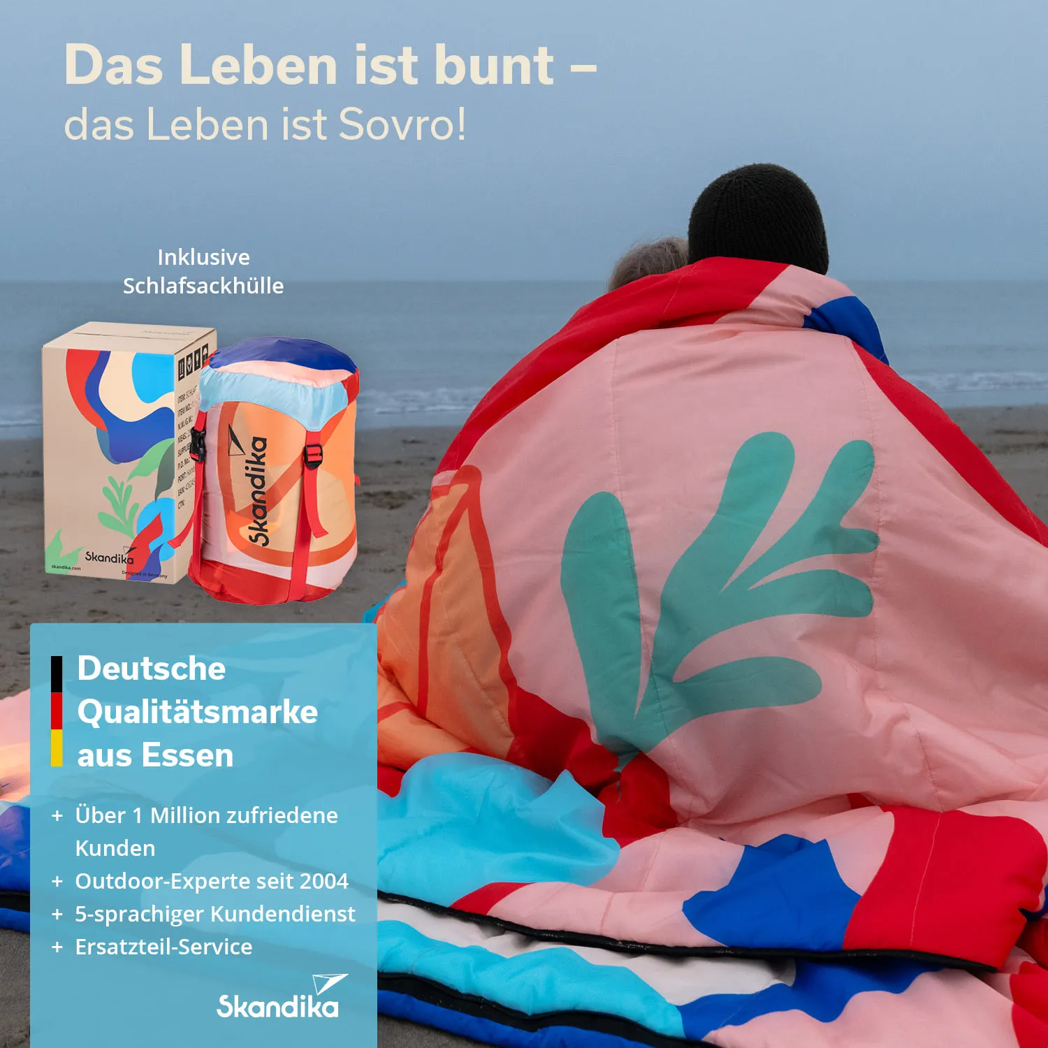 Bunter Schlafsack und Hülle auf einem Sandstrand. Im Hintergrund zwei Personen sitzen und betrachten das Meer.