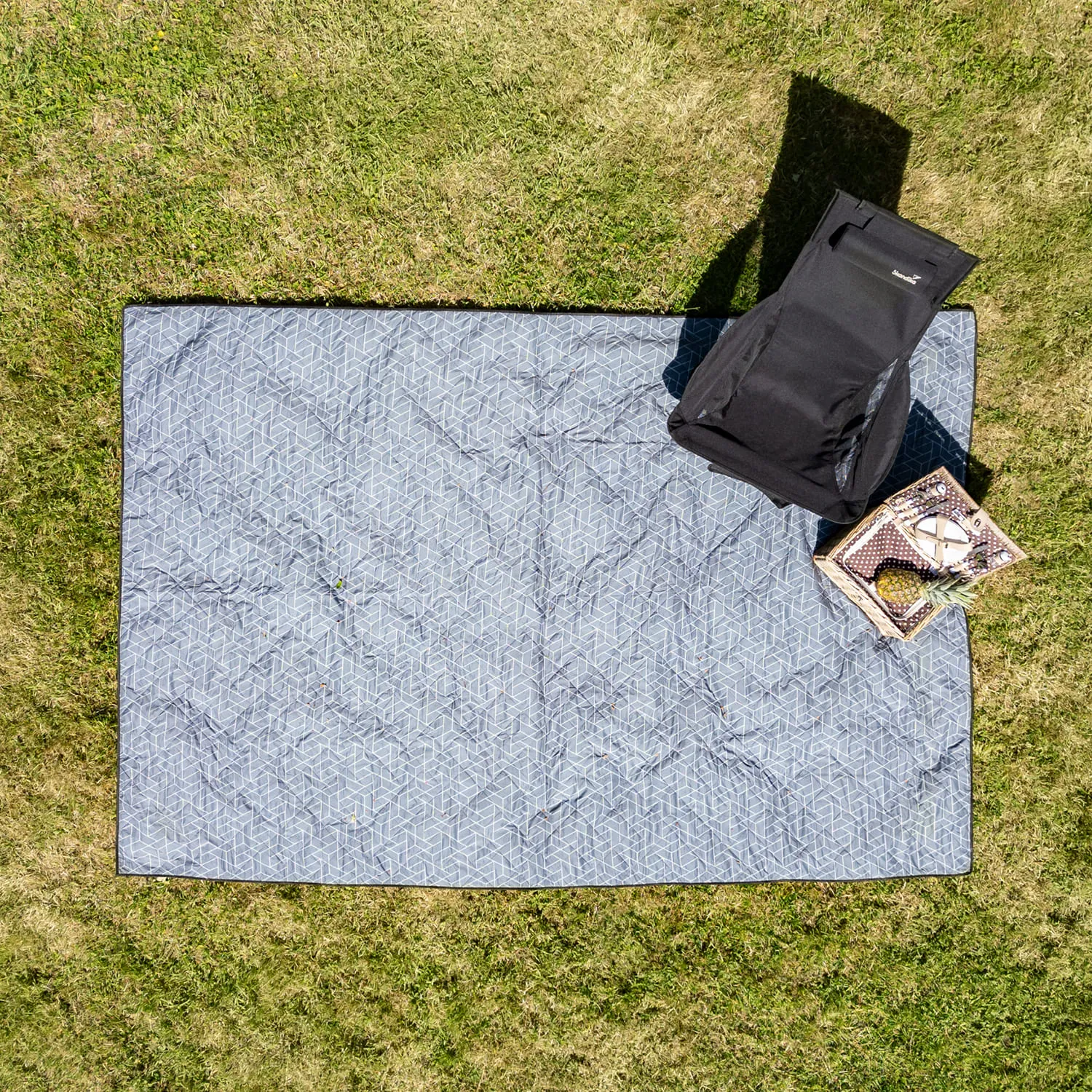 Picknickdecke mit geometrischem Muster auf grünem Gras, daneben ein schwarzer Rucksack und ein Korb mit Obst.