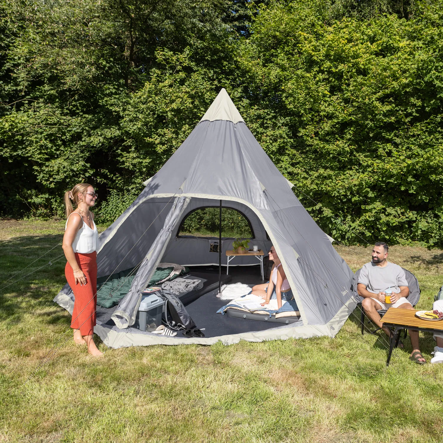 Zelt in Form eines Tipis, Personen sitzen am Tisch und stehen in der Nähe, umgeben von Grün und Wiese.