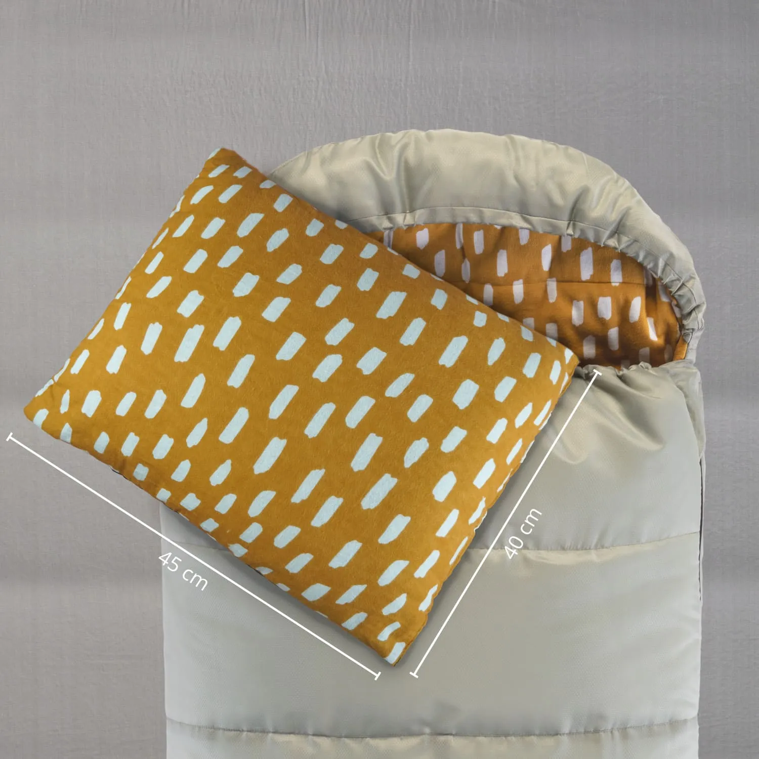 Eine beige Decke mit einem gepunkteten Inneren liegt zusammengefaltet. Ein orangefarbenes Kissen ist darauf platziert, mit blauen Muster. Maße sind angegeben.