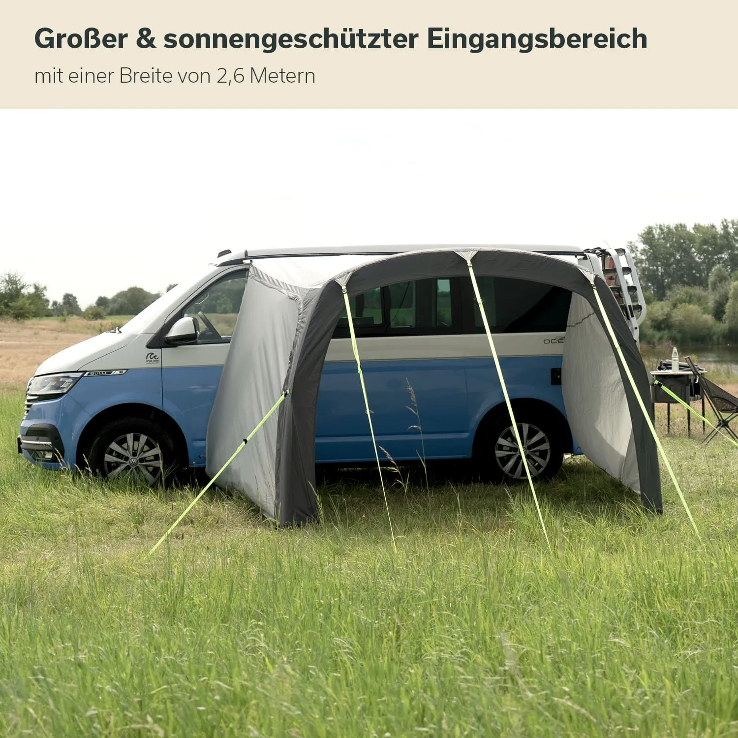 Ein blauer Campingbus steht auf einer Wiese. Vor dem Bus befindet sich ein graues, aufgestelltes Zelt mit Stangen. Im Hintergrund ist ein Feld und Bäume sichtbar.