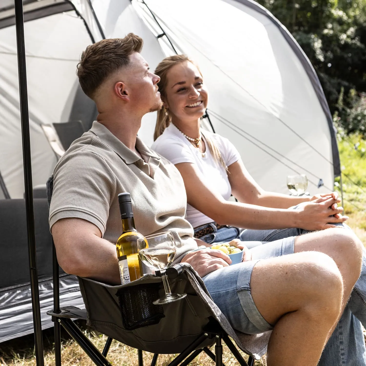 Zwei Personen sitzen in Campingstühlen vor einem Zelt. Der Mann hält eine Flasche Wein und ein Glas, die Frau hat einen kleinen Behälter mit Snacks.