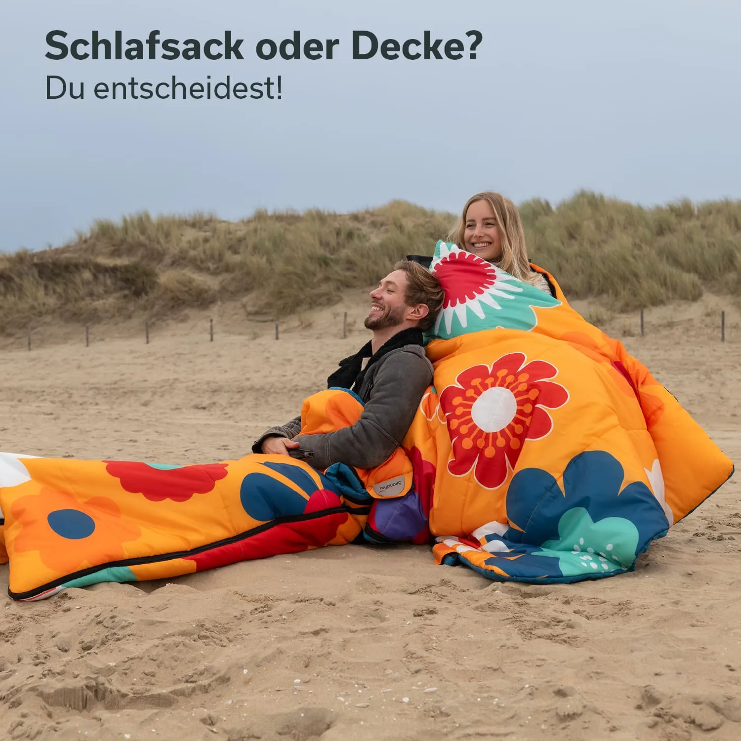 Zwei Personen sitzen im Sand, in bunte Schlafsäcke oder Decken gehüllt. Im Hintergrund sind hohe Gräser zu sehen.