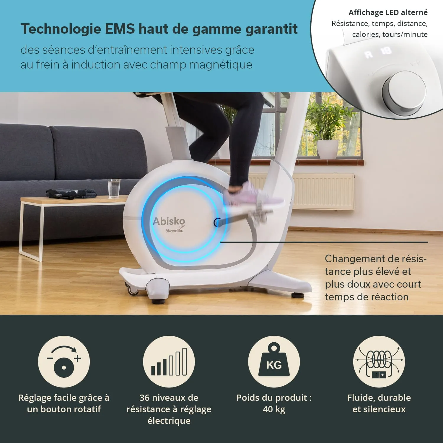 Blick auf ein Heimtrainingsgerät, einen Ergometer, mit einer Person, die darauf sitzt und strampelt. Das Gerät hat ein modernes Design mit LED-Anzeigen. Im Hintergrund sieht man ein Sofa und Fenster.