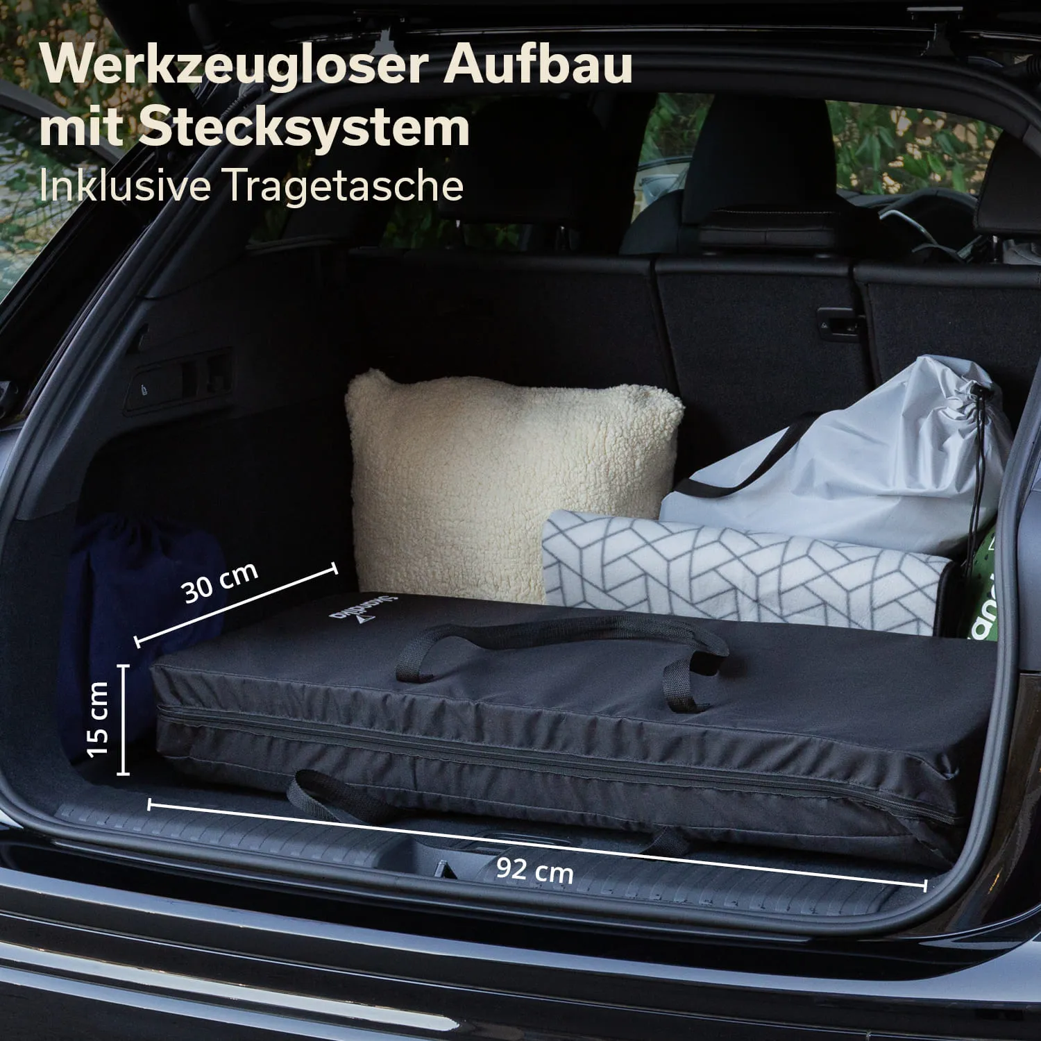 Kofferraum eines Autos mit einer schwarzen Tasche, einem Kissen, einer Decke und einer Tragetasche. Maße sind eingezeichnet.