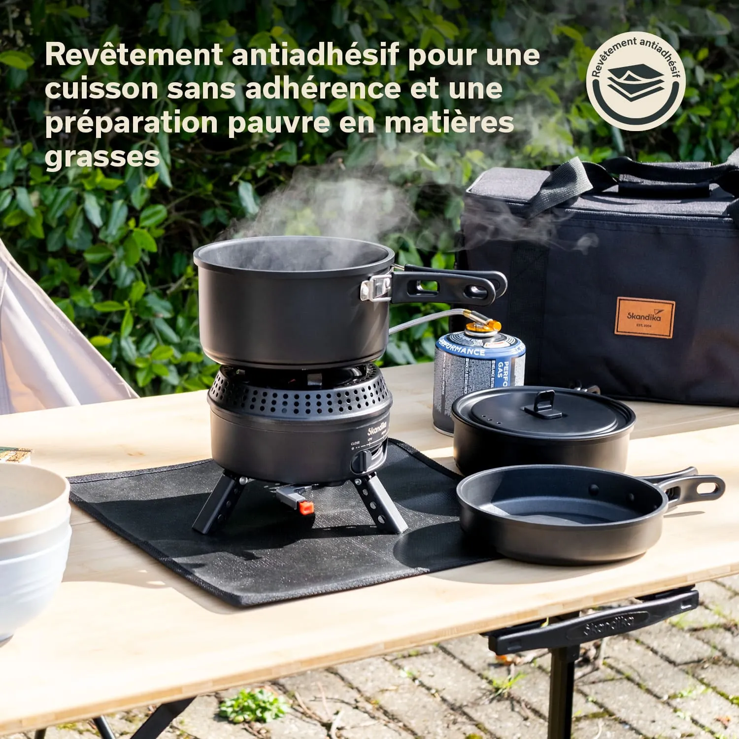 Ein mobiler Gaskocher mit einem schwarzen Topf und zwei weiteren schwarzen Kochgeräten auf einem Tisch. Dampfformation aus dem Topf.