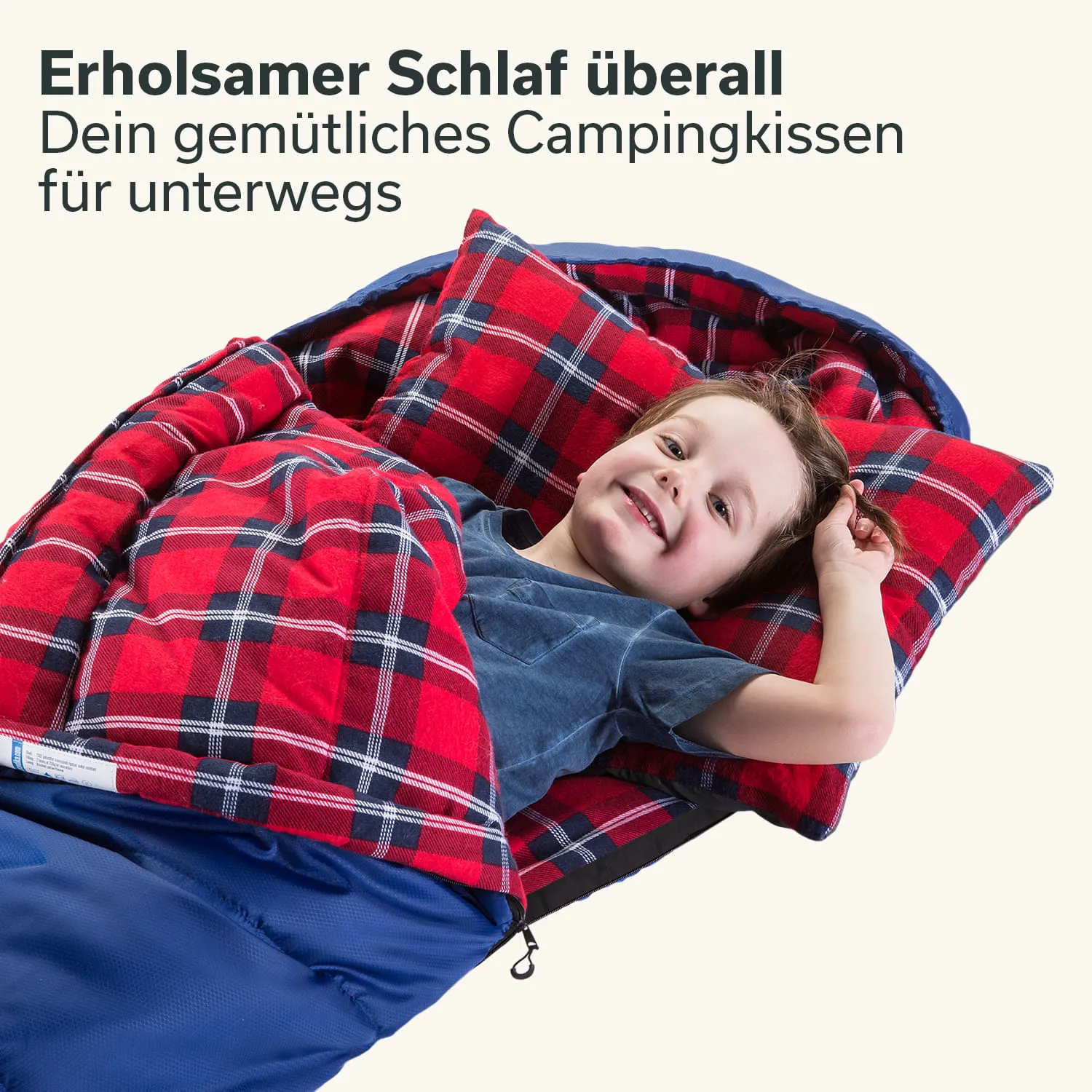 Kind liegt lächelnd in einem Schlafsack mit kariertem roten Schlafsack und Kissen. Hintergrund einfarbig.