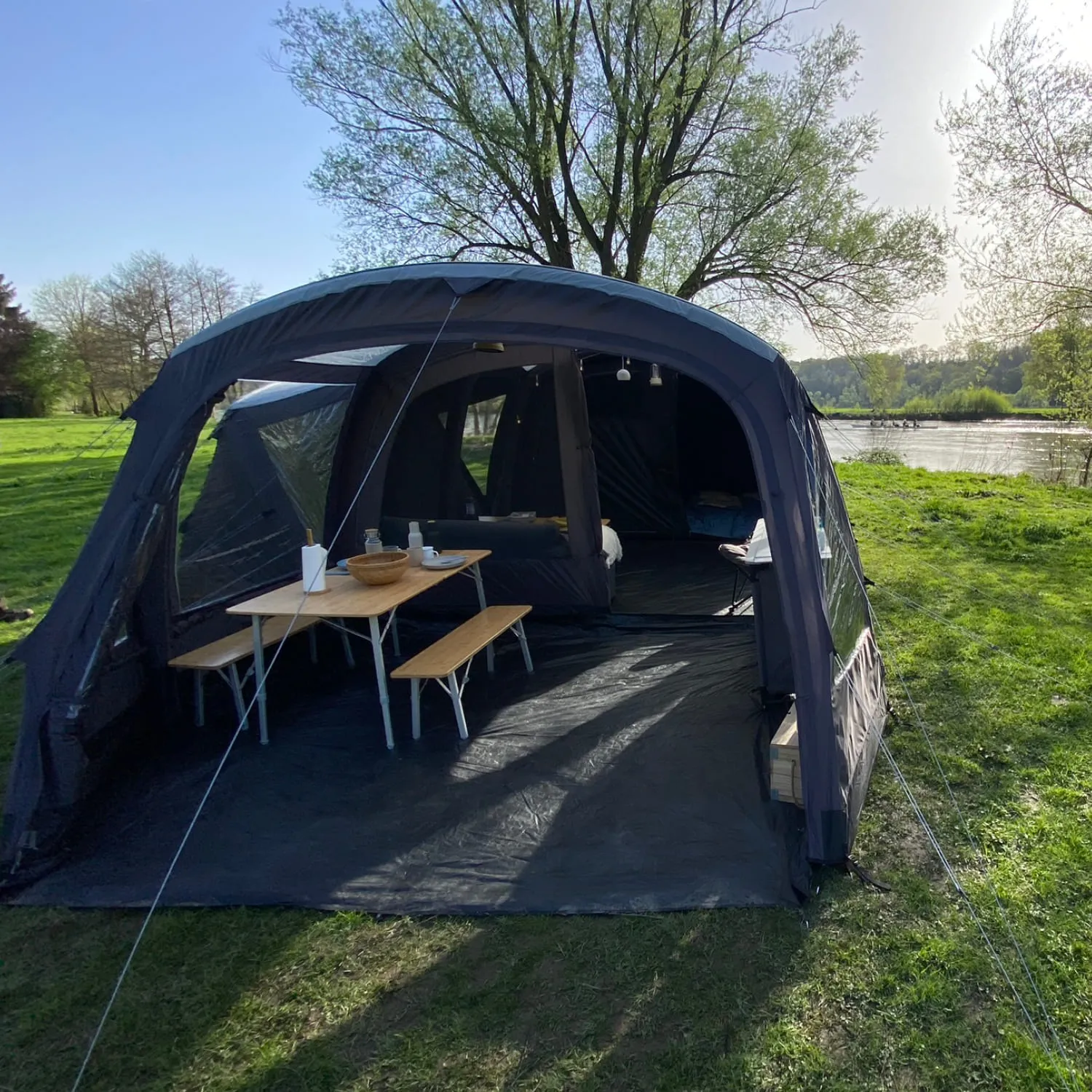 Zelt mit Tisch und Bänken auf einer Wiese, im Hintergrund ein Fluss und Bäume. Klare, sonnige Atmosphäre.