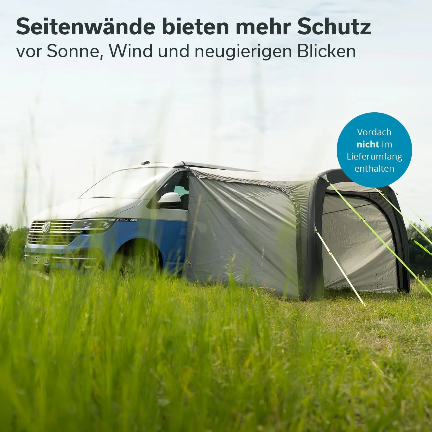 Ein blauer VW-Bus steht auf einer Wiese, an dem Seitenwände und ein Überdach befestigt sind. Hohe Gräser umgeben das Fahrzeug.