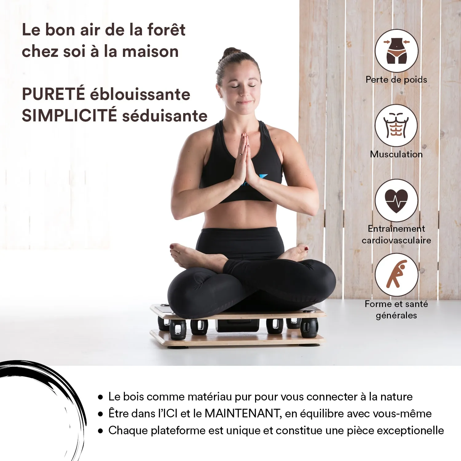 Eine Person sitzt im Schneidersitz auf einer Plattform, hält die Hände vor der Brust in einer Meditationshaltung. Die Person trägt sportliche Kleidung. Im Hintergrund sind helles Holz und Fenster zu sehen.