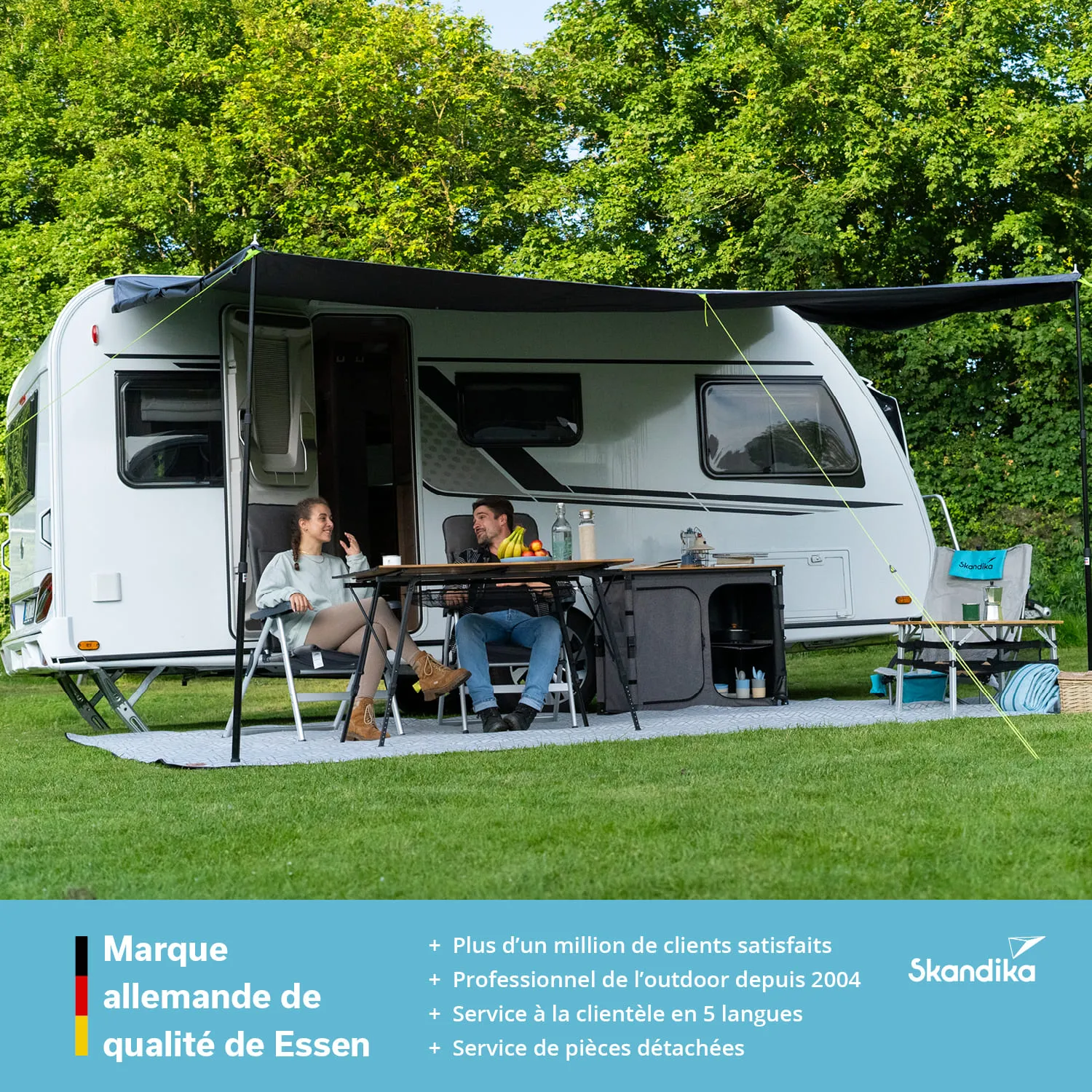 Caravan auf einer Wiese mit einer Sitzecke und einem Tisch. Zwei Personen sitzen entspannt am Tisch. Grüne Bäume im Hintergrund.