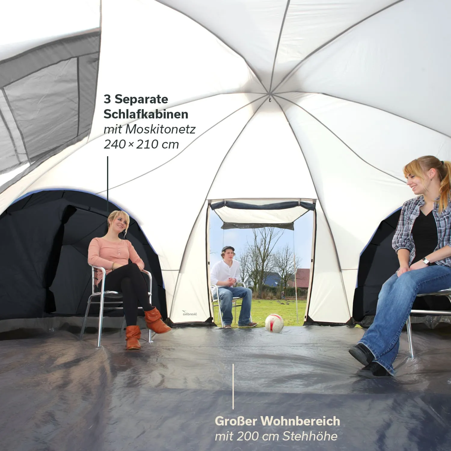Großes Zelt mit drei Schlafkabinen, innen sitzen drei Personen, darunter ein Fußball, kahle Bäume im Hintergrund.
