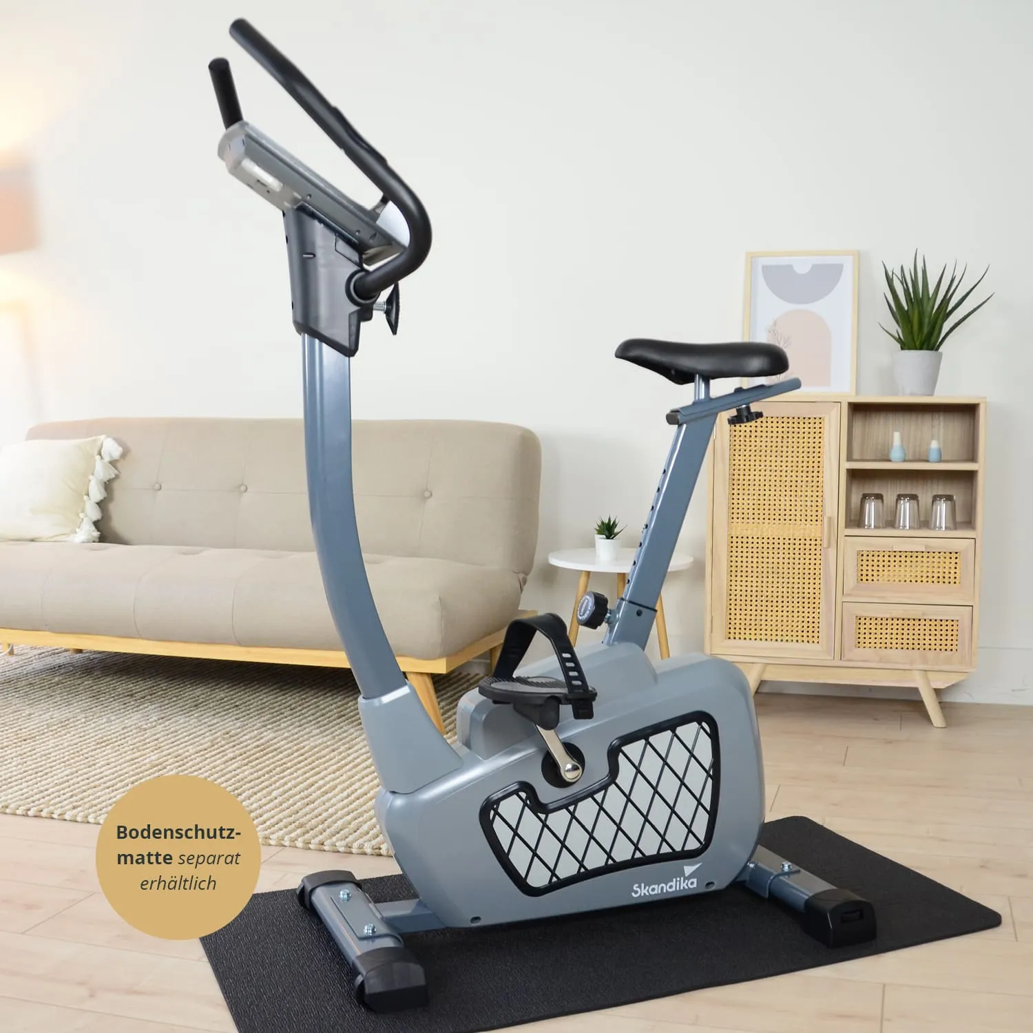Ergometer in grauer Farbe auf einer Bodenmatte, im Hintergrund eine Couch, ein Beistelltisch und ein Regal mit Pflanzen.
