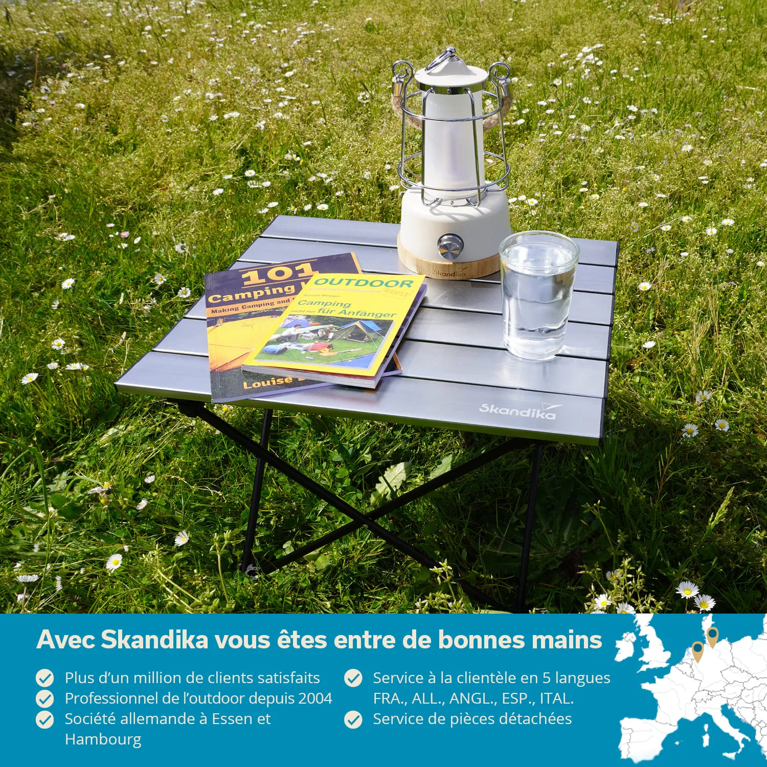 Klappbarer Tisch aus Aluminium mit zwei Campingbüchern, einer Lampe und einem Glas Wasser auf einer Wiese.