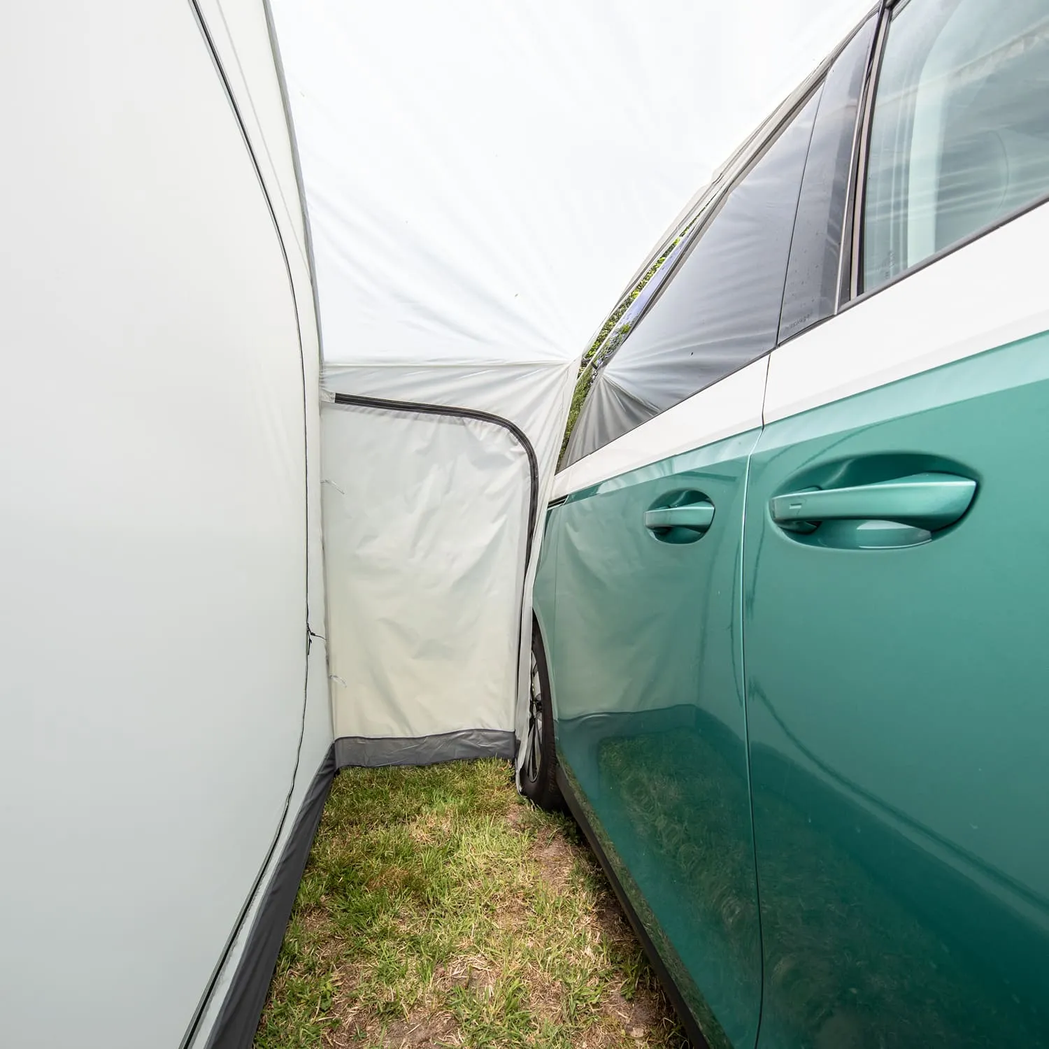 Ein grünes Auto steht neben einer weißen Zeltwand. Der Zelteingang ist geöffnet und führt nach außen. Grass ist sichtbar.