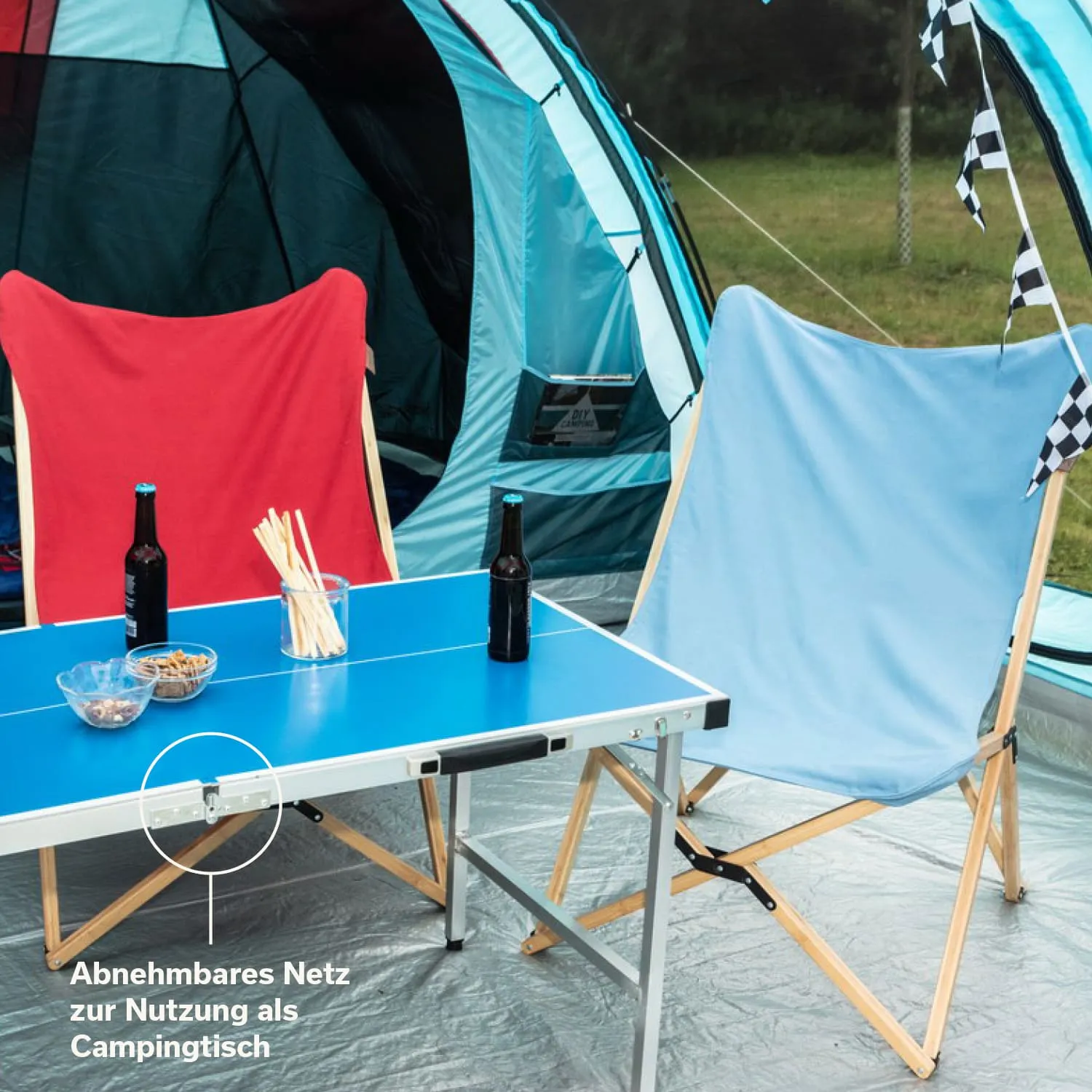 Zwei Stühle, rot und blau, stehen an einem blauen Tisch. Darüber sind zwei Flaschen und Snacks. Im Hintergrund ein Zelt und Wiese.