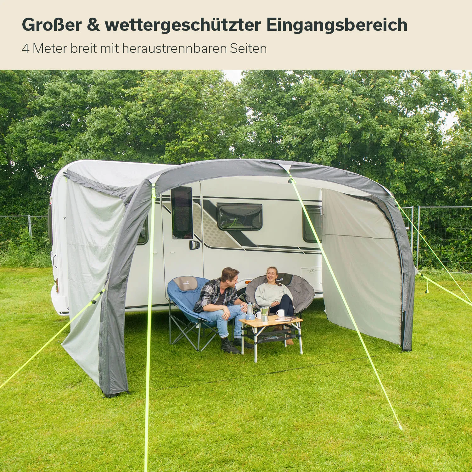 Stellplatz mit Wohnwagen und einem überdachten Bereich. Zwei Personen sitzen auf Campingstühlen und trinken Kaffee.