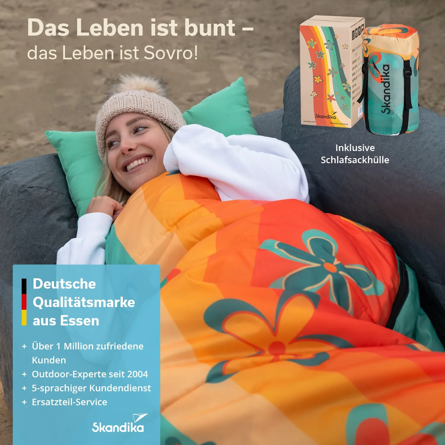 Eine Person liegt lächelnd in einem bunten Schlafsack mit Blumenmuster auf einer Couch. Im Hintergrund sind Kissen sichtbar und ein Produktkarton.