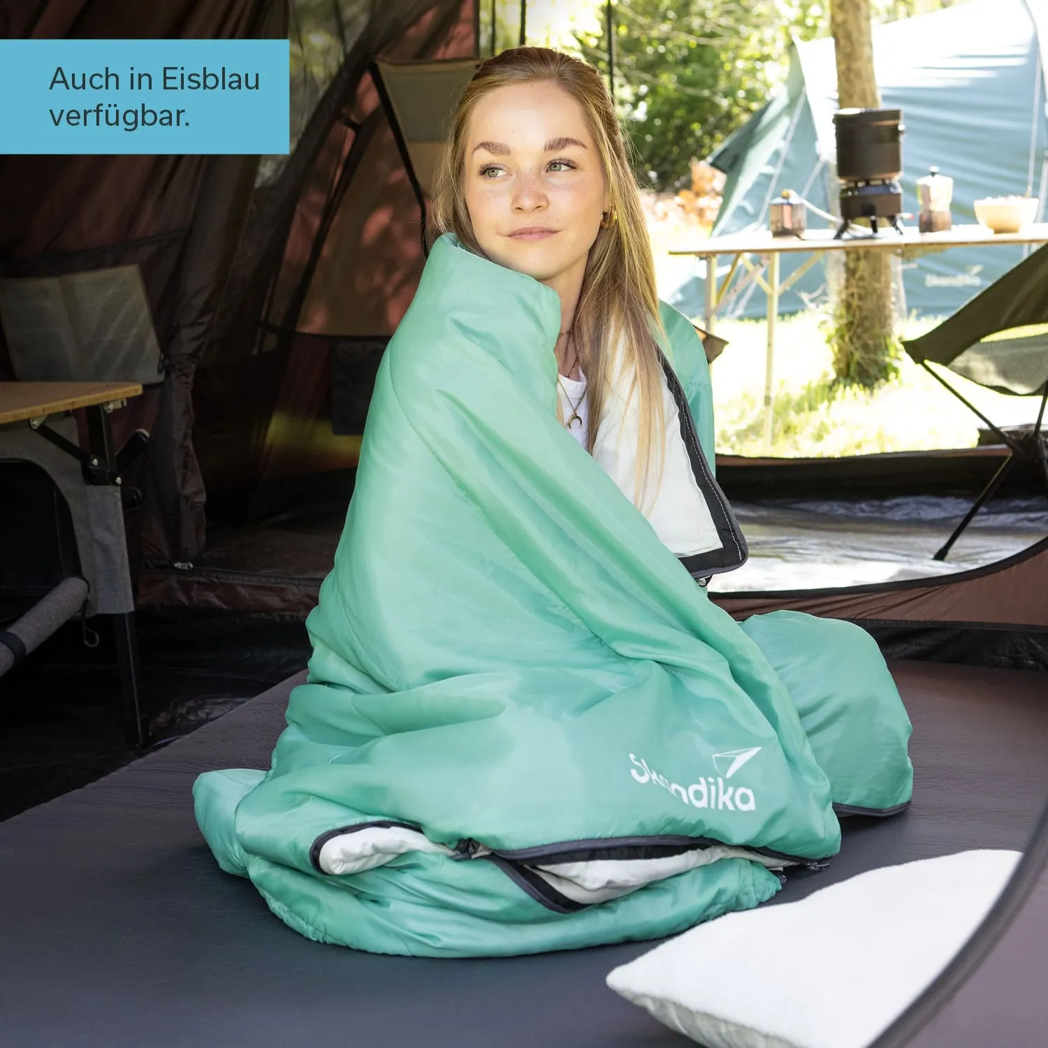 Eine Person sitzt auf einer Matratze in einem Zelt, eingehüllt in einen mintgrünen Schlafsack. Im Hintergrund sind Zelte und ein Tisch sichtbar.